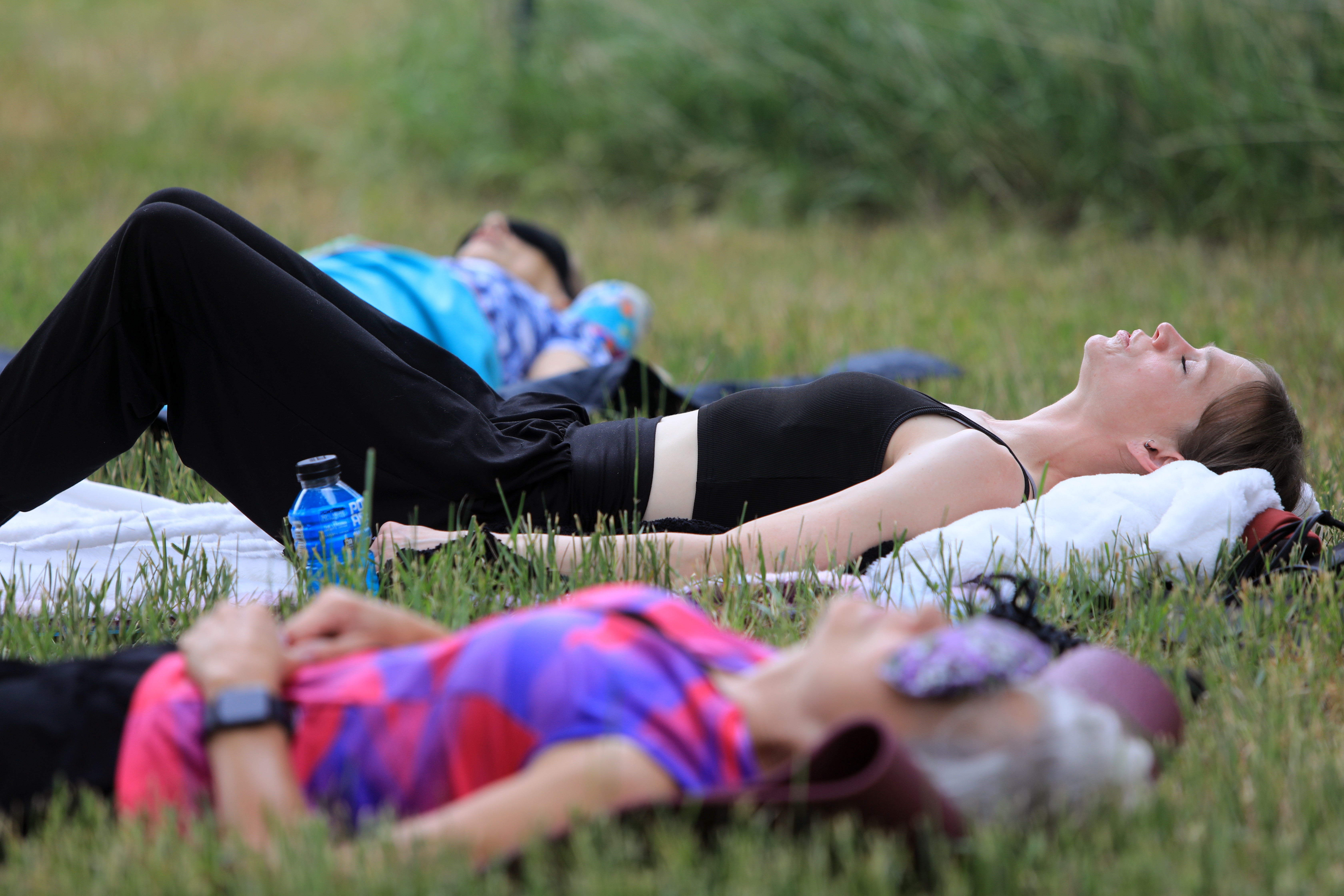 Two people lying on grass outdoors, one in the foreground wearing a colorful shirt, the other in the background in black and blue, relaxing with eyes closed.