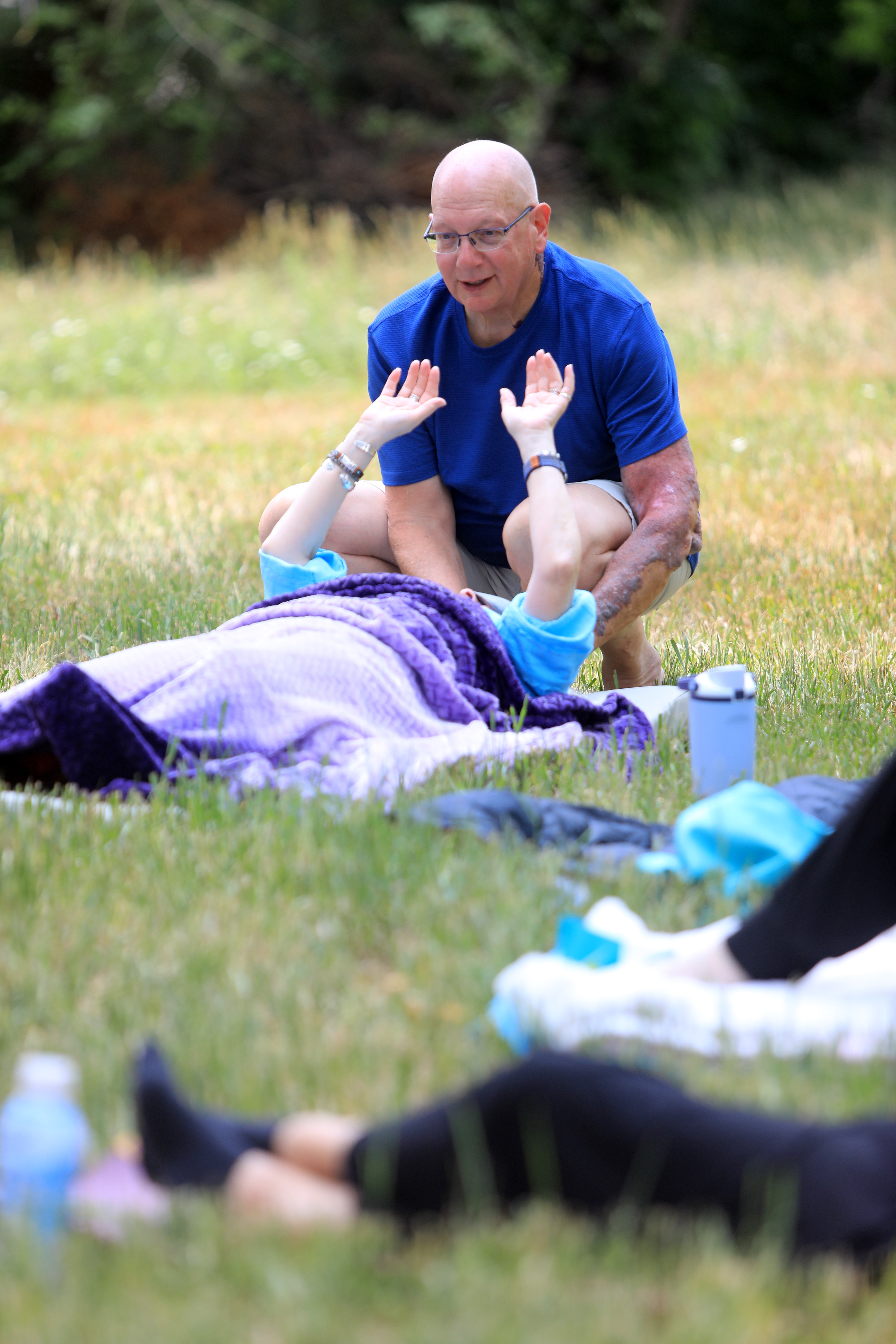 A man with glasses and a blue shirt is kneeling outdoors, smiling at a child lying on a blanket, raising hands.