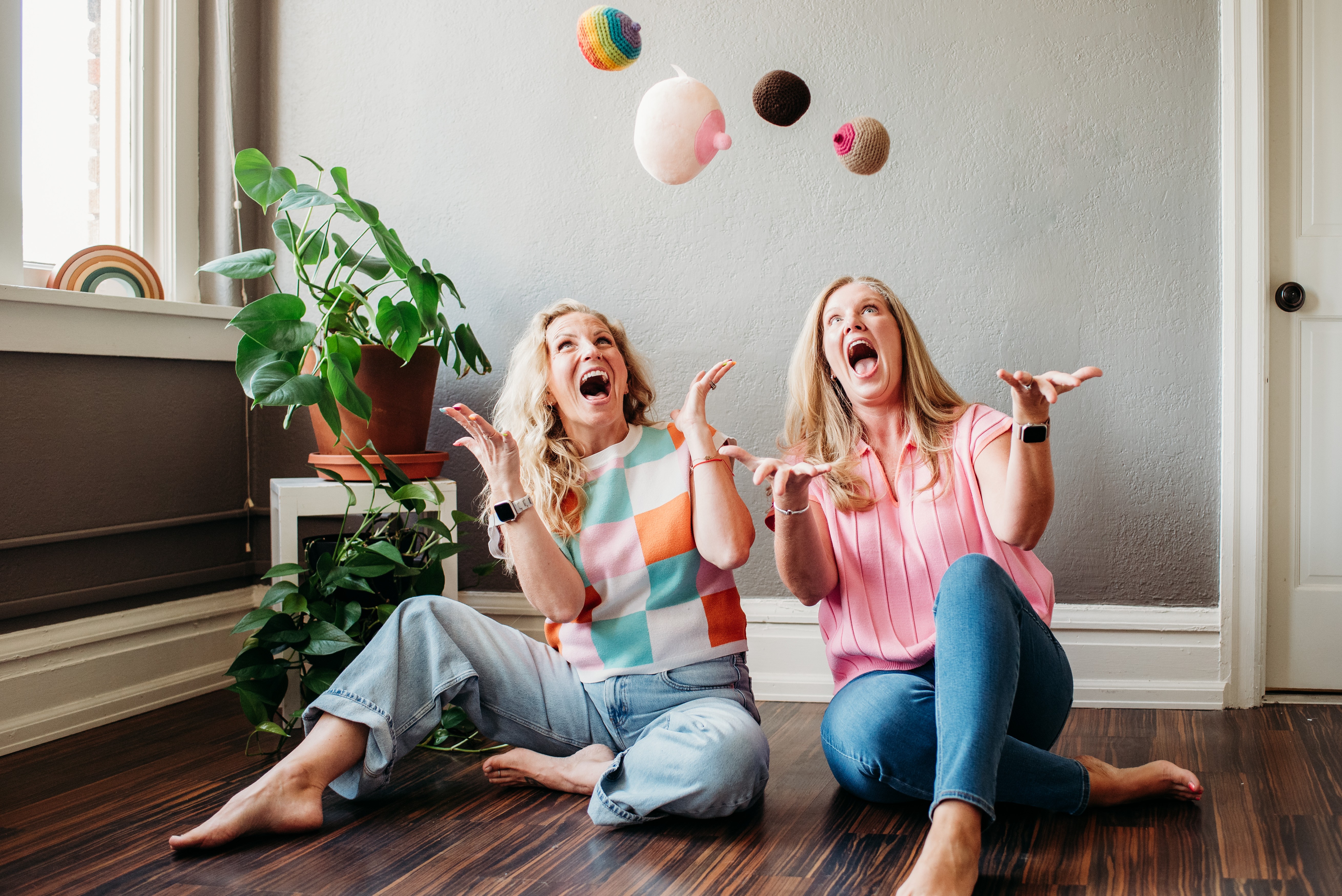 Two women sitting on a wooden floor, laughing and playing with colorful balls in a room with a potted plant and window, smiling.