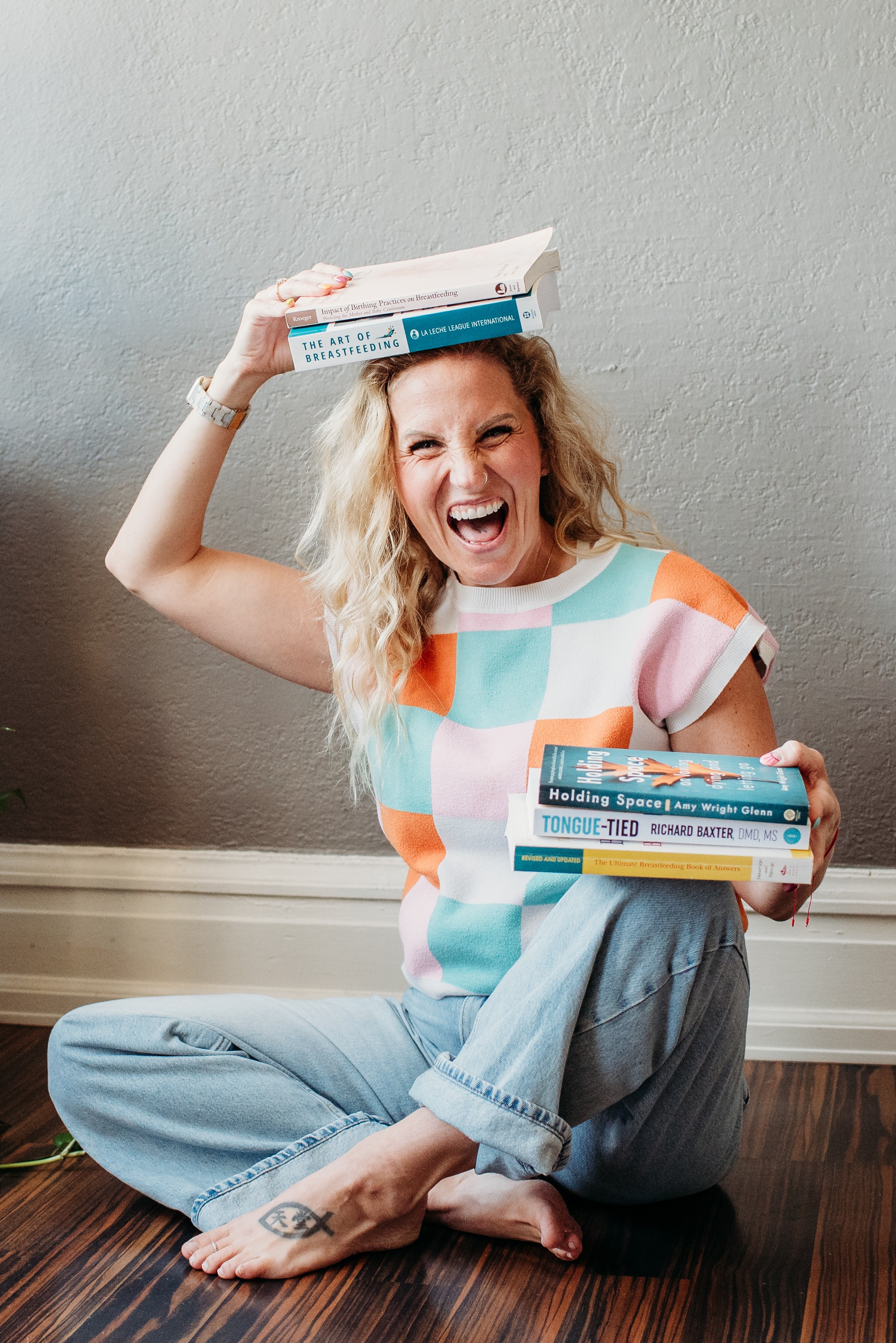 Woman sitting on floor, holding books, smiling, wearing a colorful striped shirt and jeans, against a plain wall.