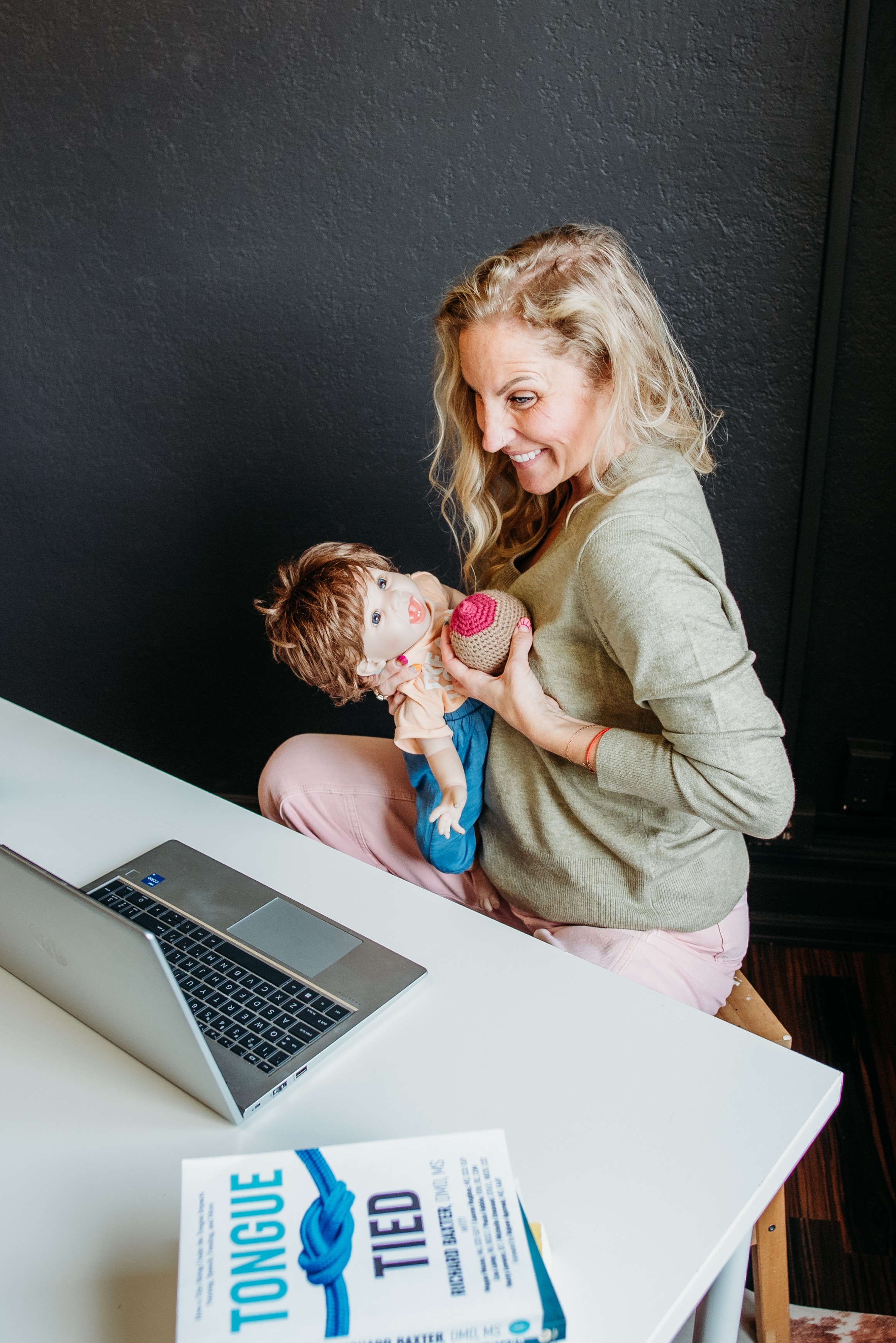 Woman holding a baby girl with a pink toy, sitting at a white table with a laptop and a brochure, smiling.