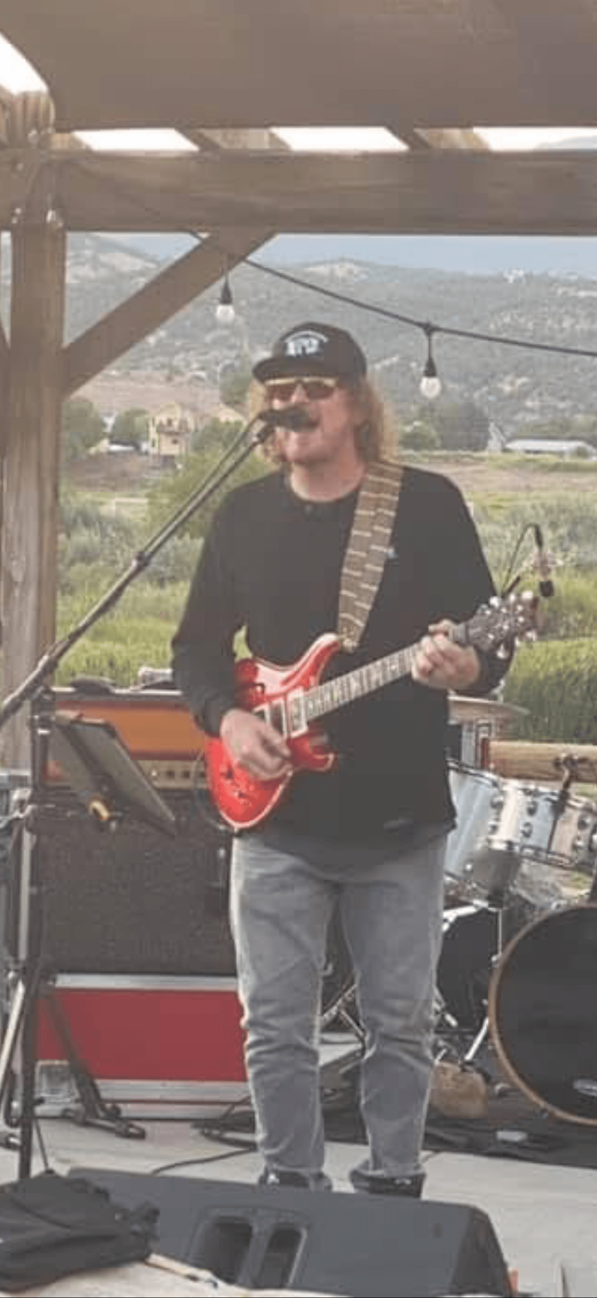 Person playing red electric guitar on outdoor stage, wearing sunglasses, black hat, black shirt, and jeans.