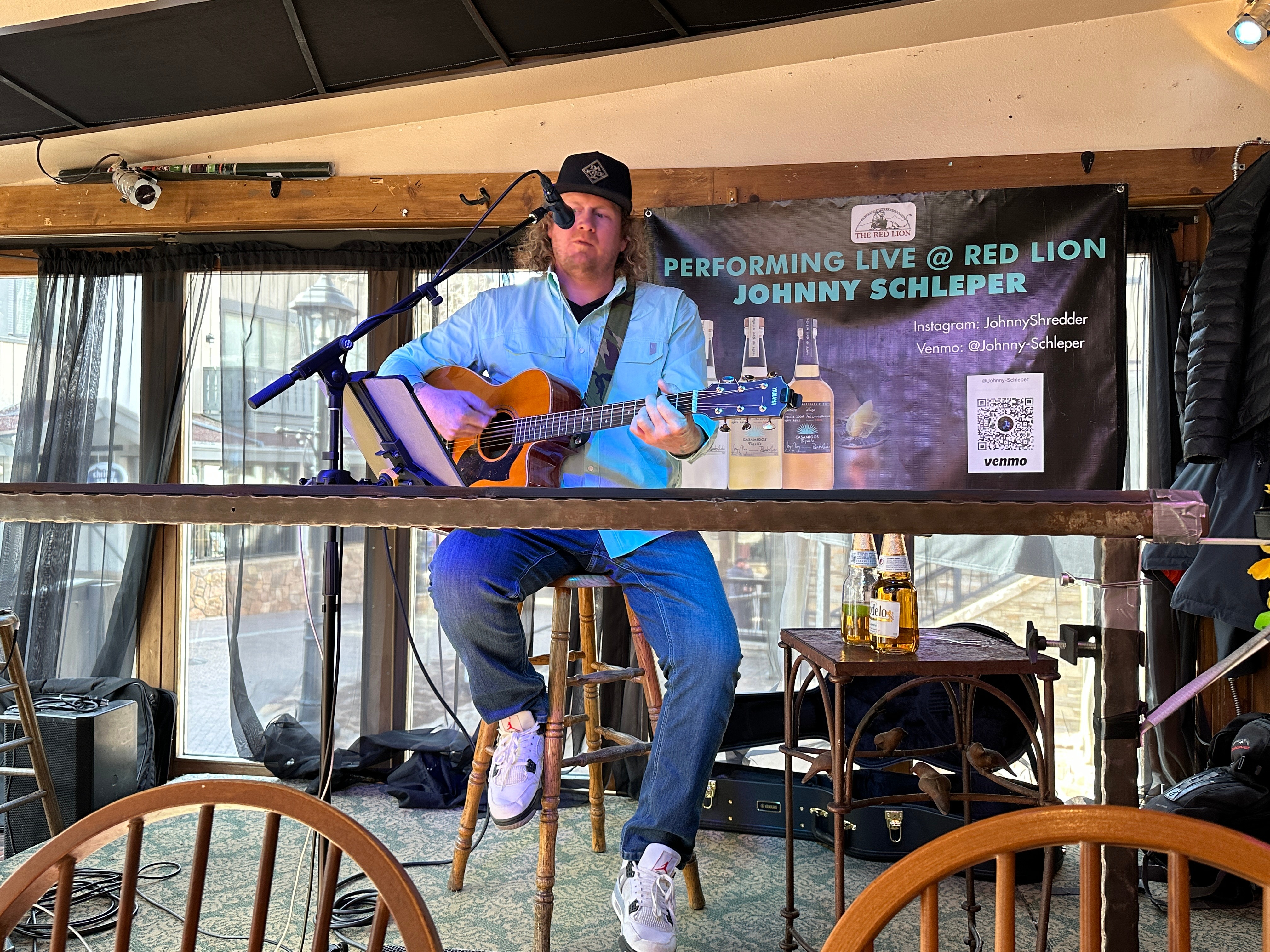 Male musician playing guitar on stage with a banner behind him, performing live at Red Lion, with a stool and bottles nearby.