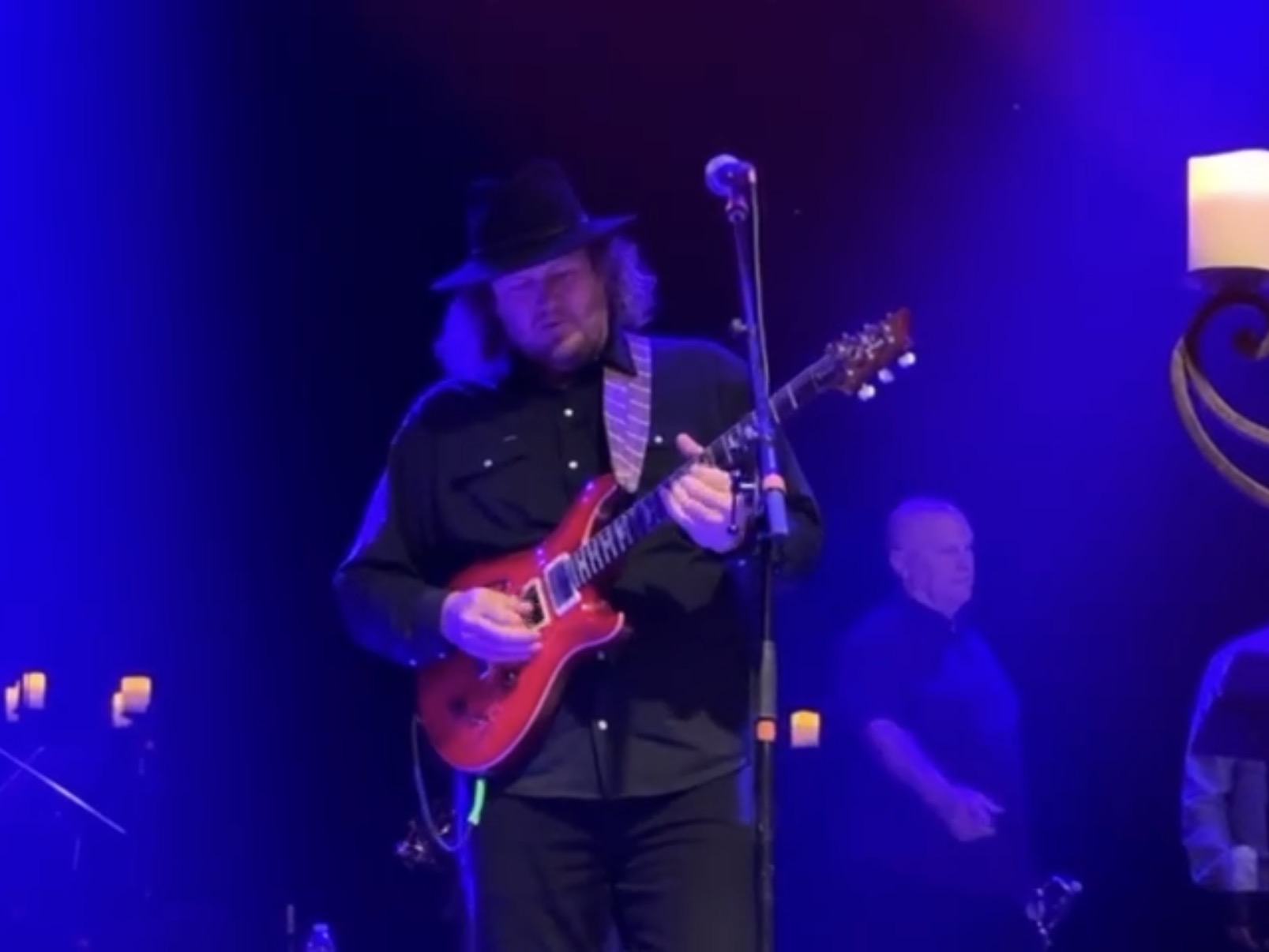 Musician with long hair, hat, and dark clothing playing a red electric guitar on stage with blue lighting.