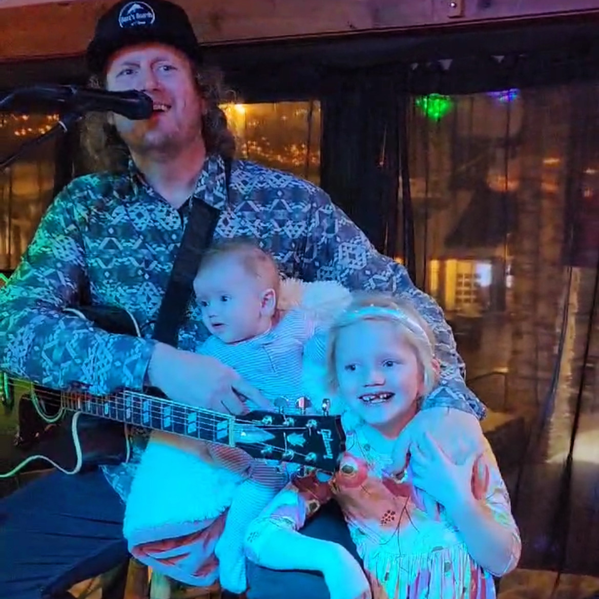 Man with guitar and two children, one girl smiling, in a cozy indoor setting with warm lighting.