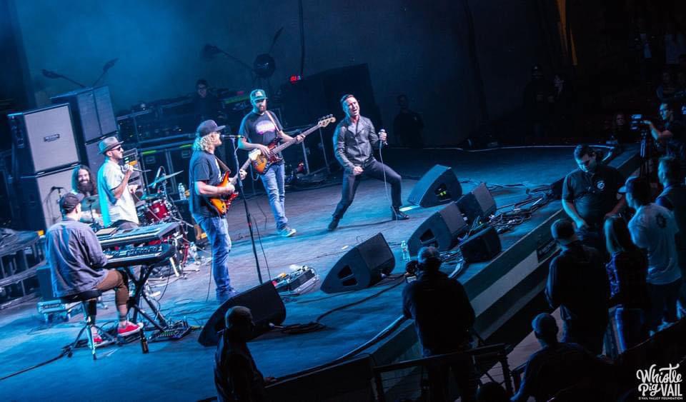 Band performs on stage with five musicians, audience watching, stage lights, and sound equipment visible.
