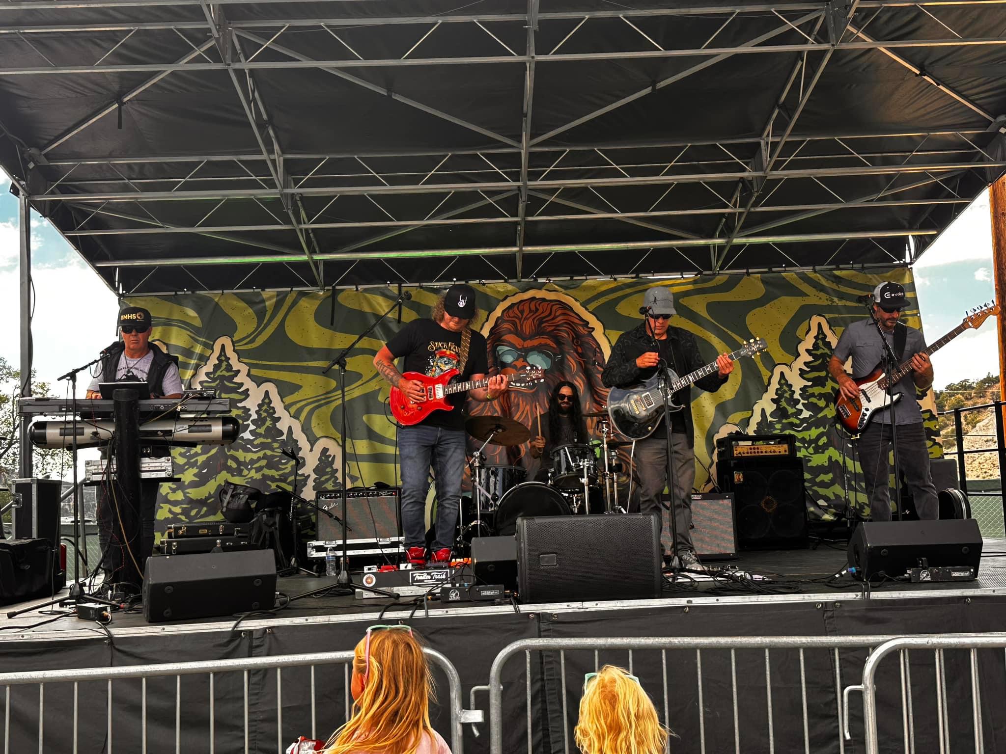 Band performing on stage with five musicians, colorful backdrop, and audience barrier in front.