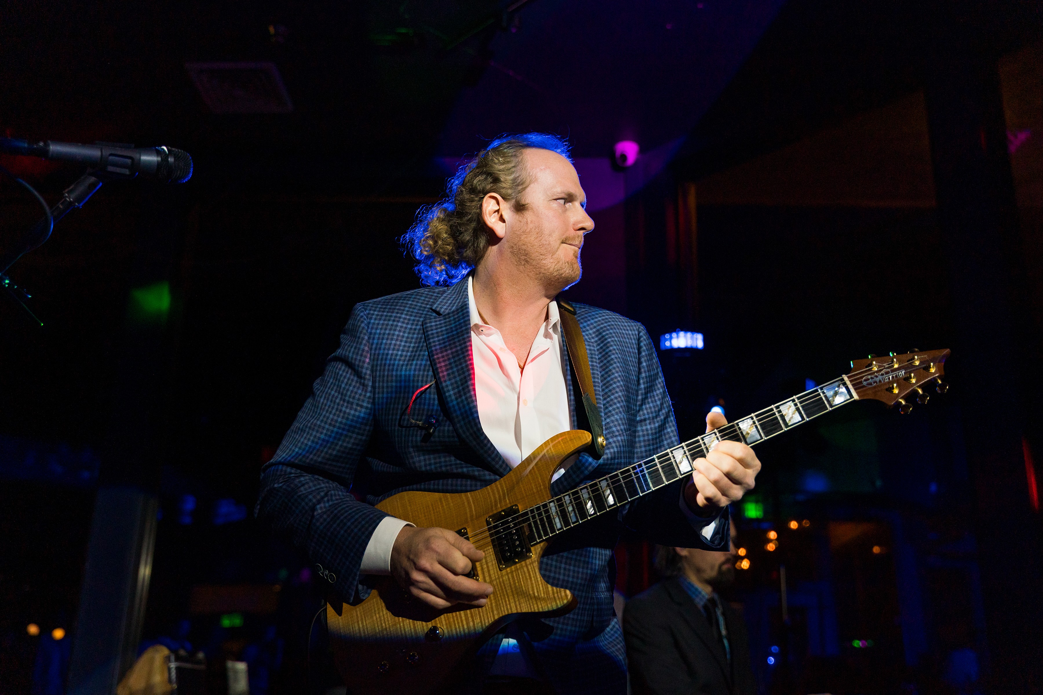 Man playing an electric guitar on stage with dark background and colorful lights, wearing a suit jacket and white shirt.