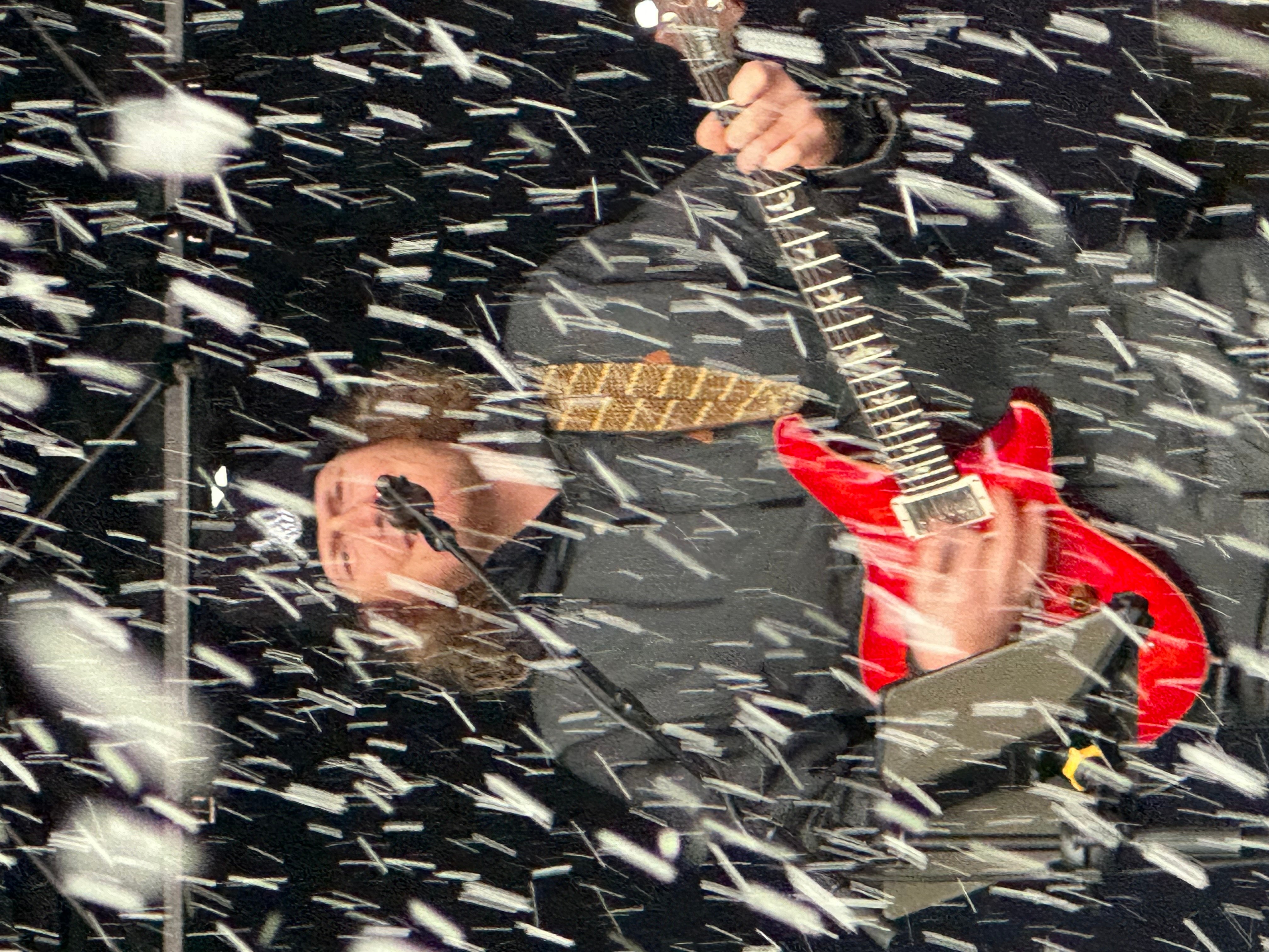 Person playing red electric guitar amidst falling confetti or snow, with a dark background.