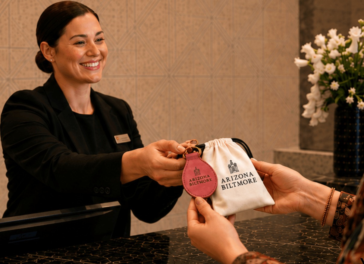 A woman in a black blazer hands a small bag to another person, with a flower arrangement in the background.