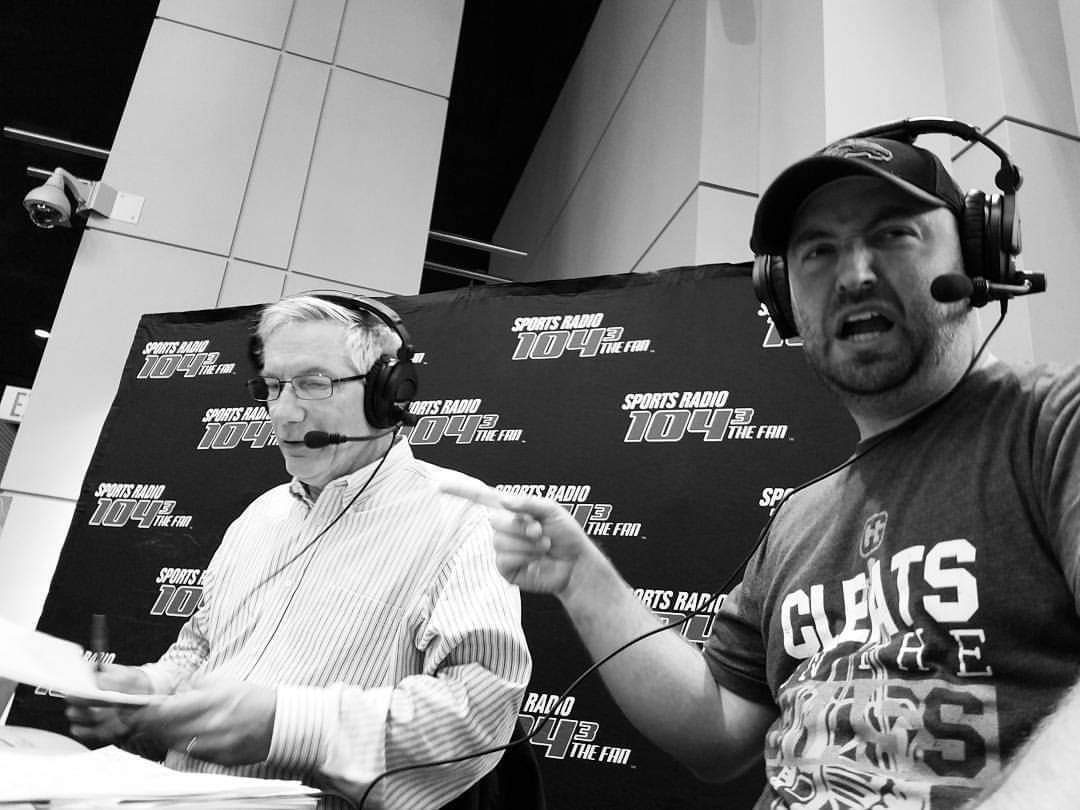 Two men wearing headsets and microphones, one in a striped shirt and glasses, speaking at a sports radio broadcast.