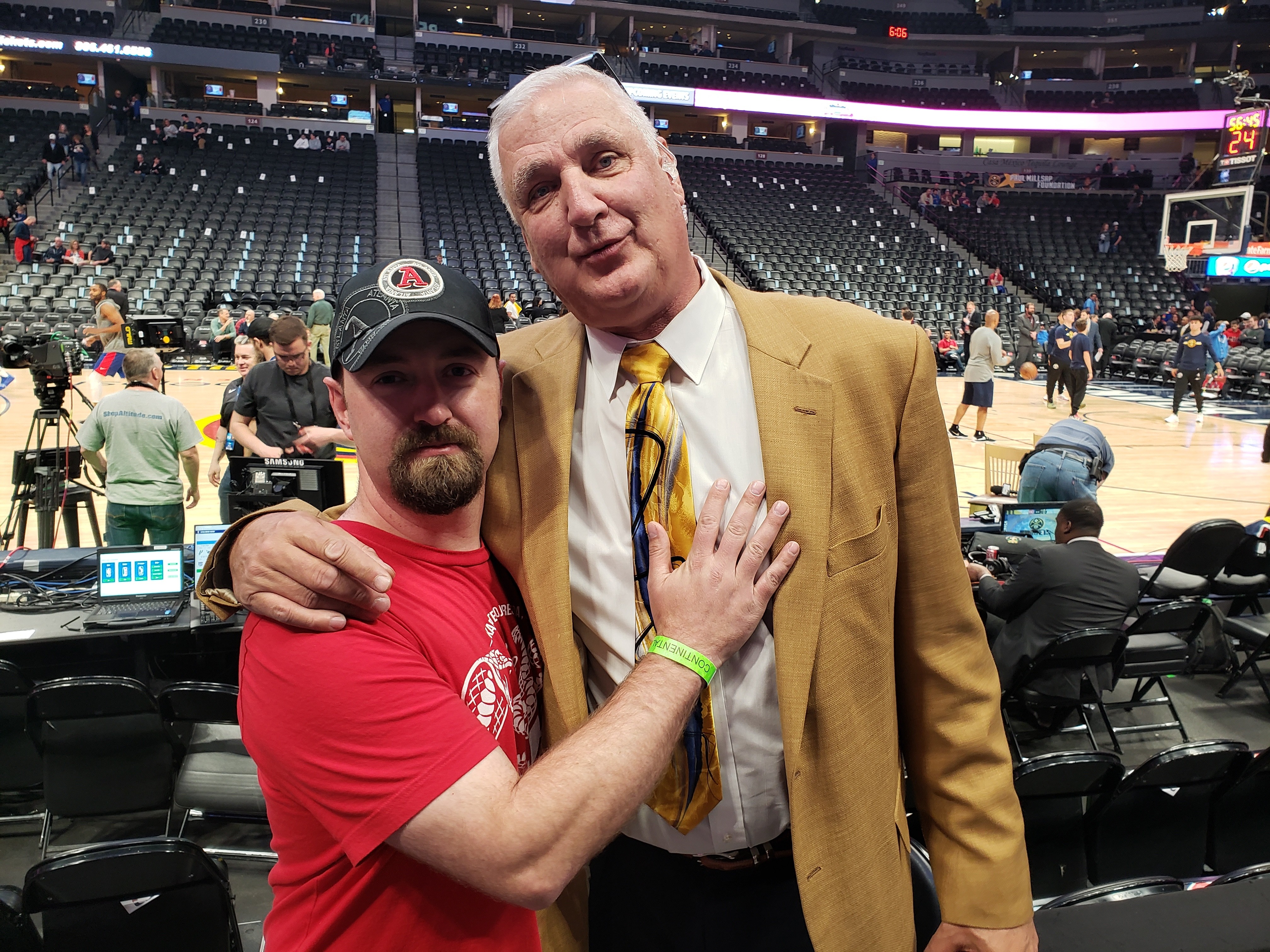 Two men standing close together in a sports arena, one in a red shirt and the other in a tan suit, with empty seats in the background.