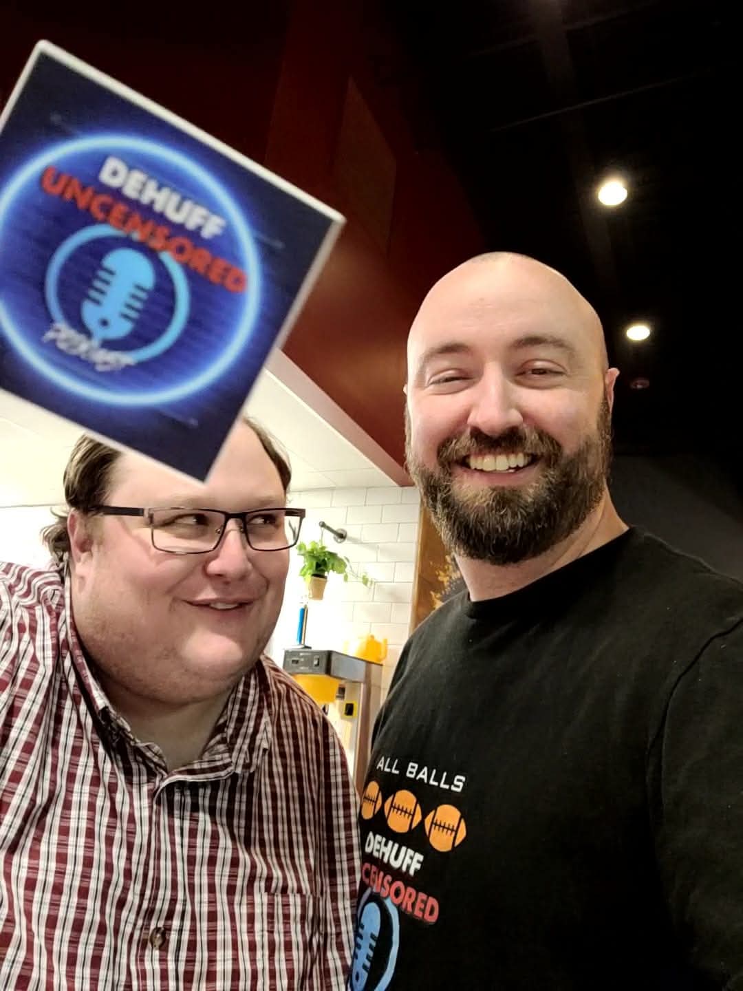 Two men smiling indoors, one wearing glasses and a checkered shirt, the other with a beard and black shirt, with a sign in the foreground.