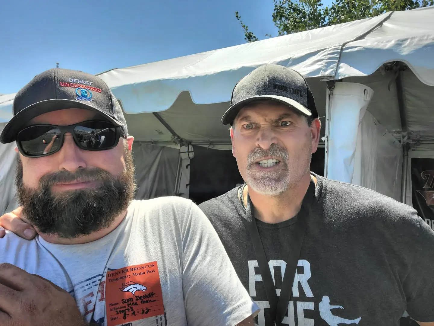 Two men outdoors, one with sunglasses and a beard, the other wearing a cap, standing in front of a white tent.