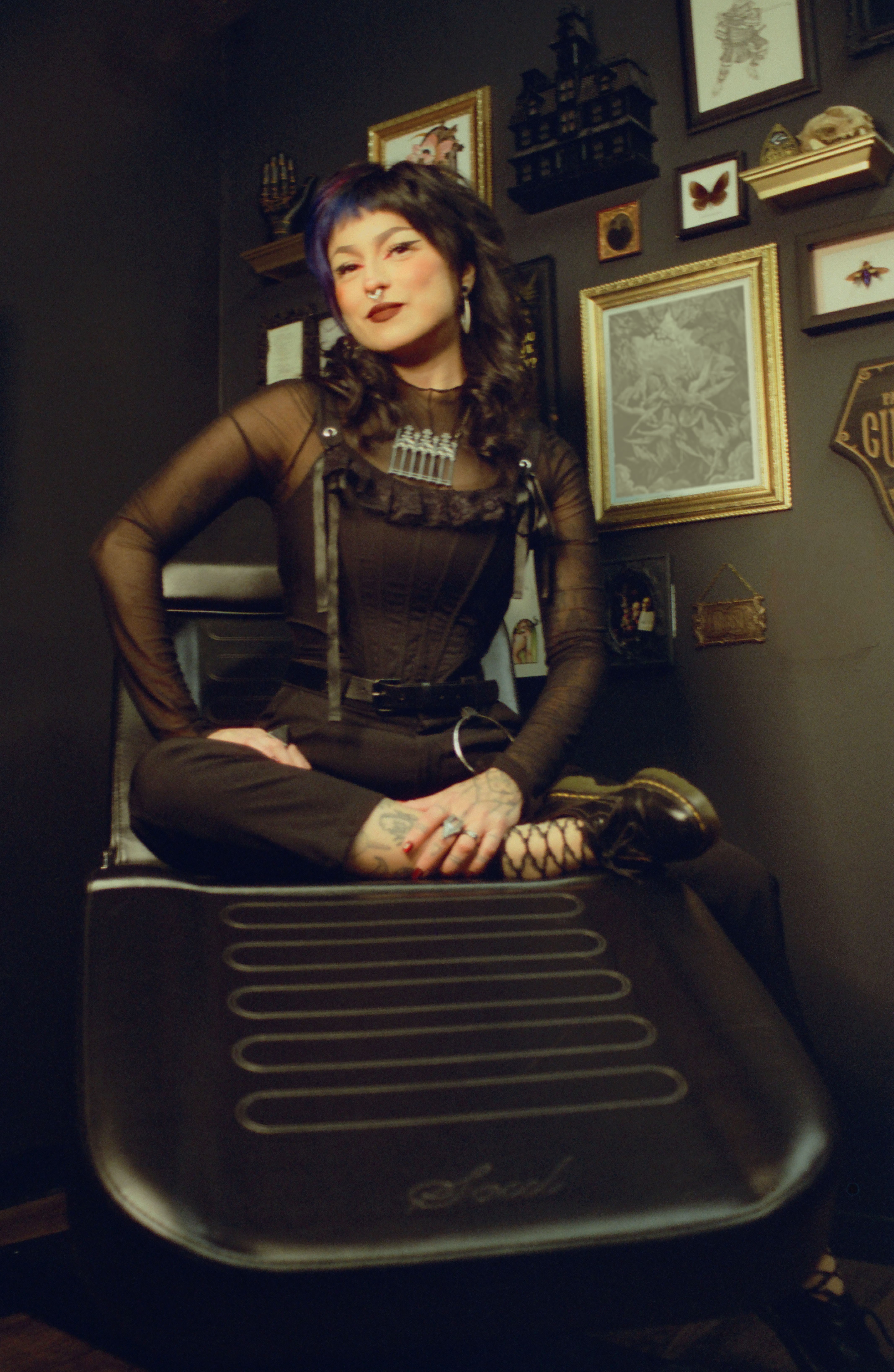 Woman with dark curly hair sitting cross-legged on a black object, surrounded by framed pictures and decorations on a dark wall.