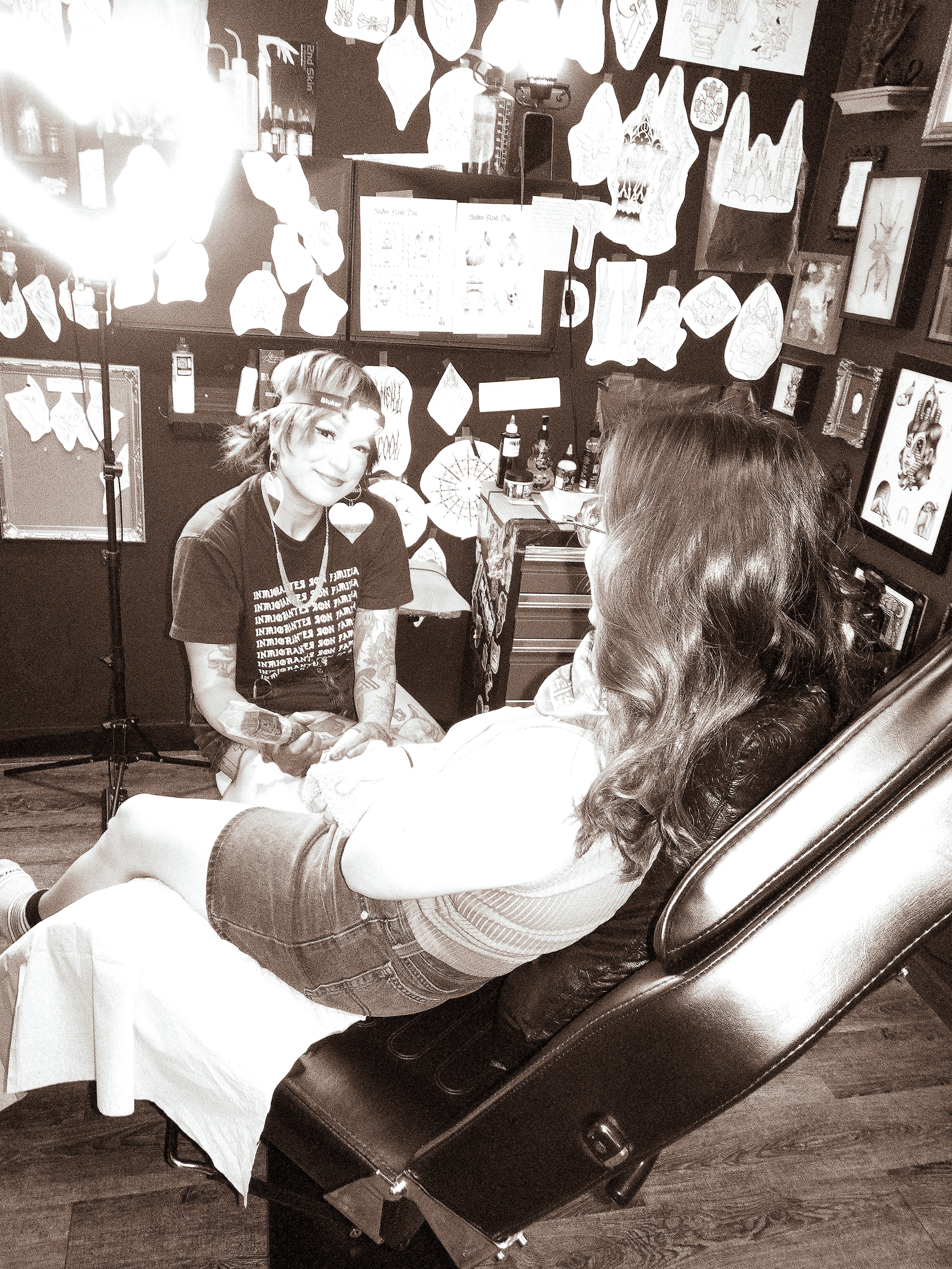 A woman with long hair is seated in a tattoo chair, talking to a tattoo artist in a studio with artwork on the walls.