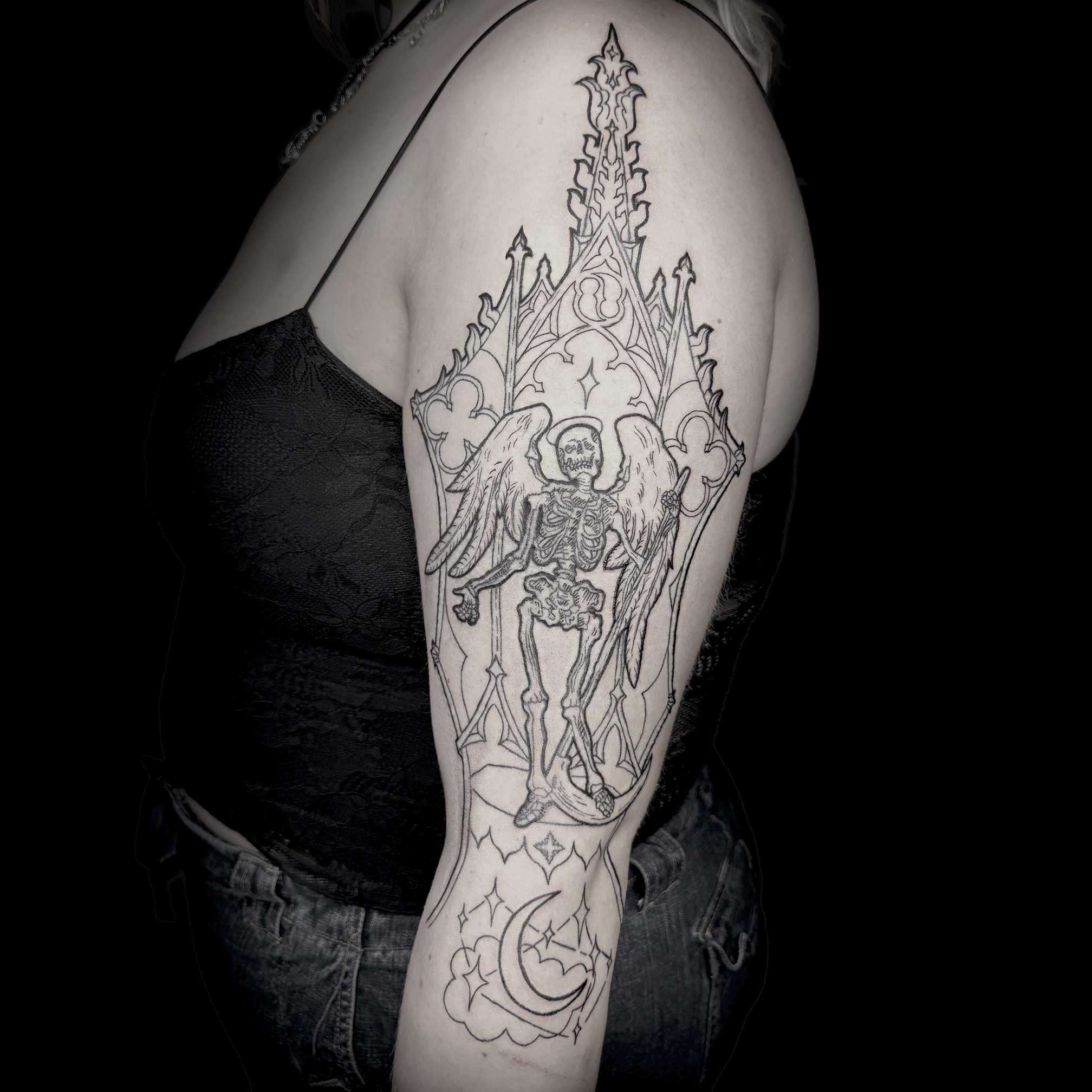 Black and gray tattoo of a winged skeleton with a sword on upper arm, moon, and decorative elements, on person's arm.