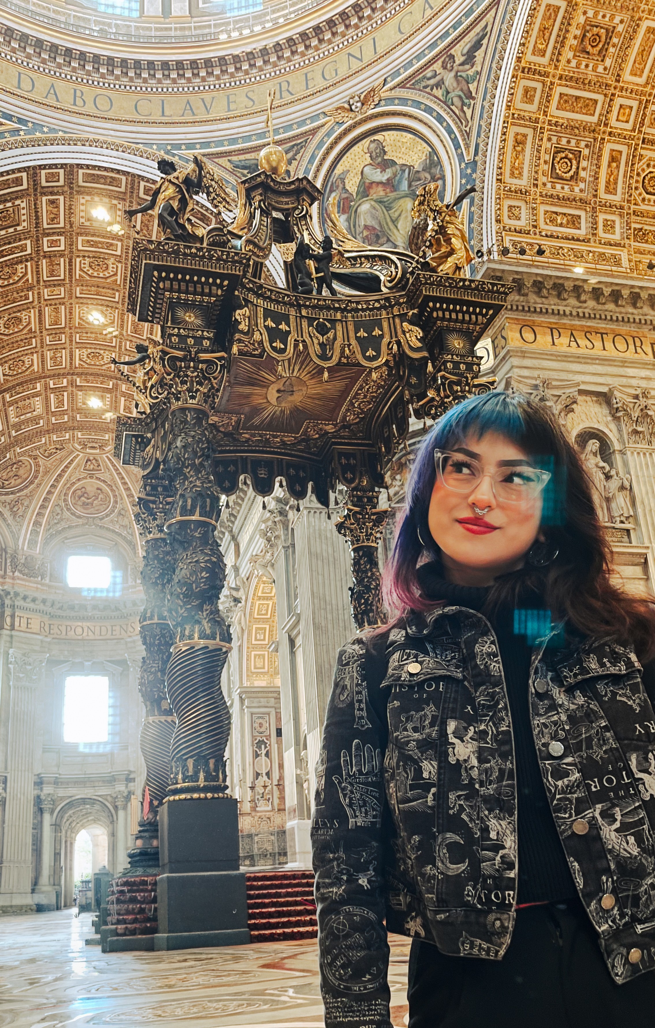 Young woman with dark hair and red lipstick standing inside a grand, ornate church with gold decorations and sculptures.