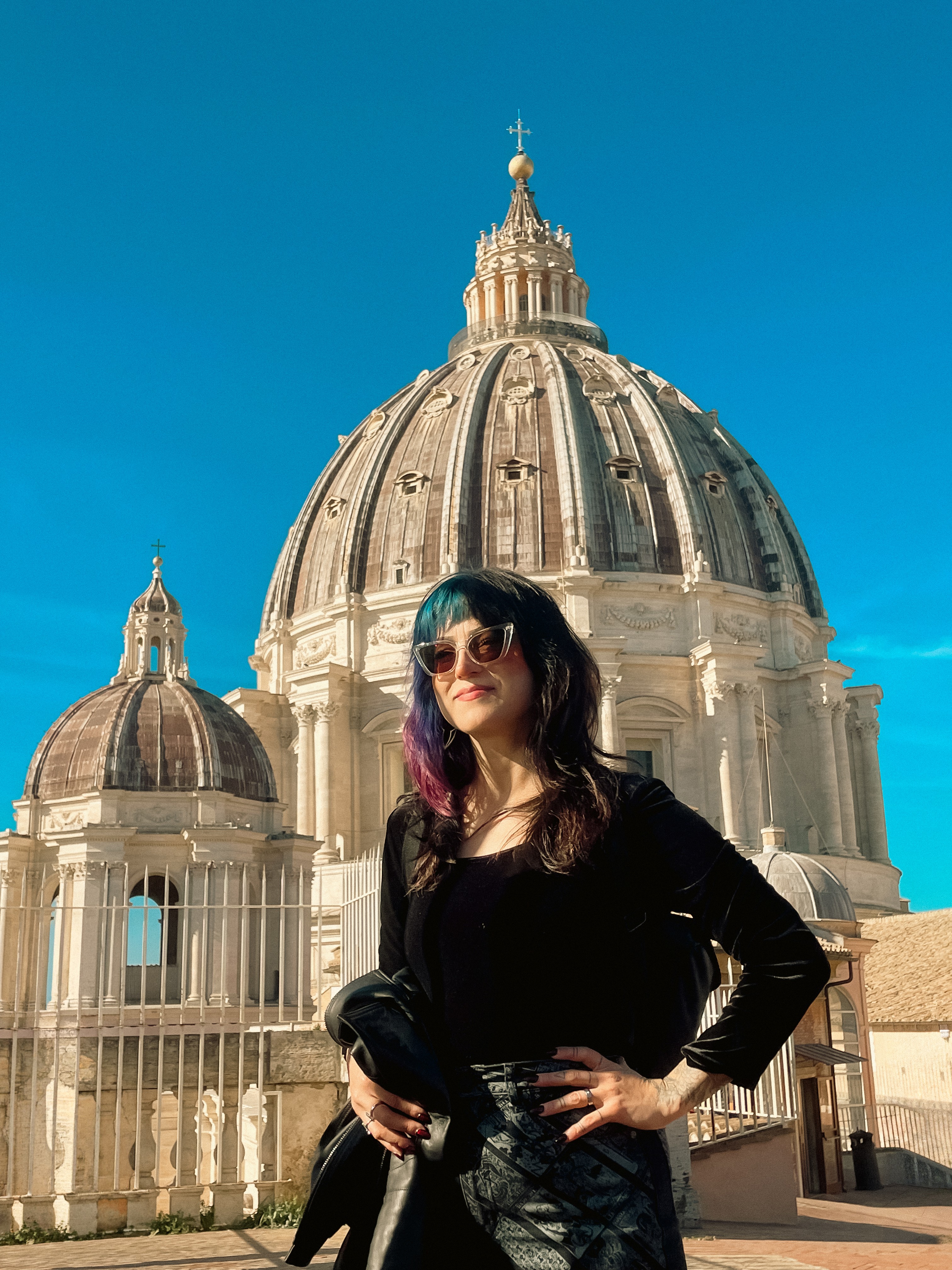 Woman with sunglasses and dark hair standing in front of a large domed building under a clear blue sky.