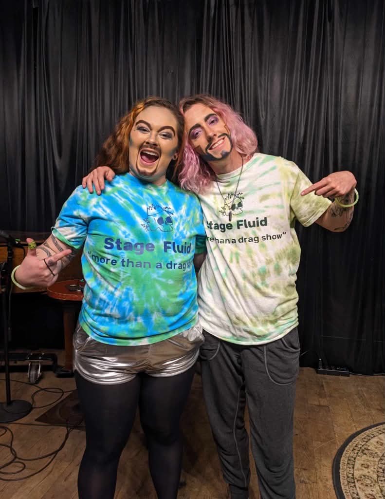 Two people smiling and posing with arms around each other in front of a black curtain, wearing tie-dye shirts with text.