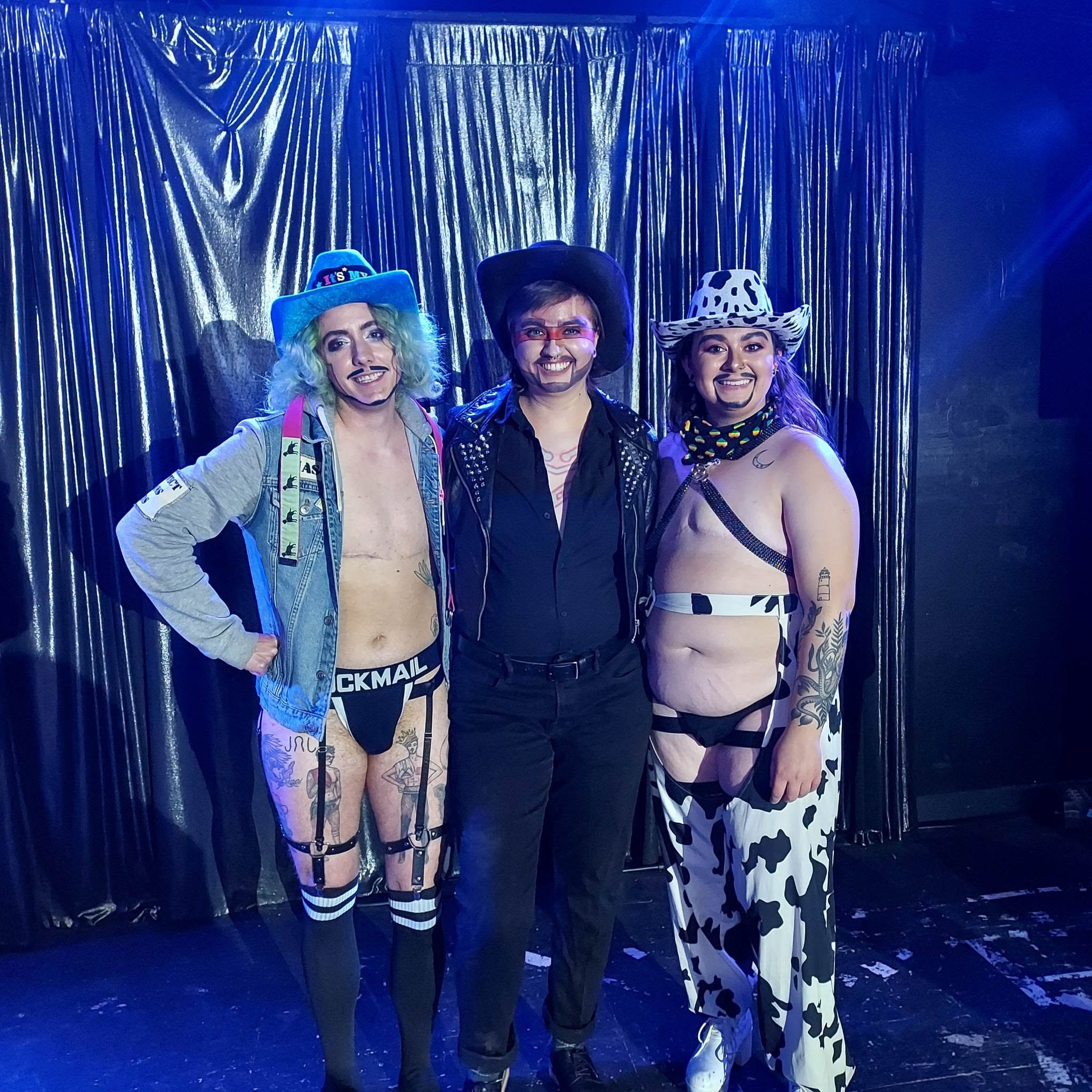Three people in costumes pose in front of a shiny curtain, two with cowgirl outfits and one in black attire.