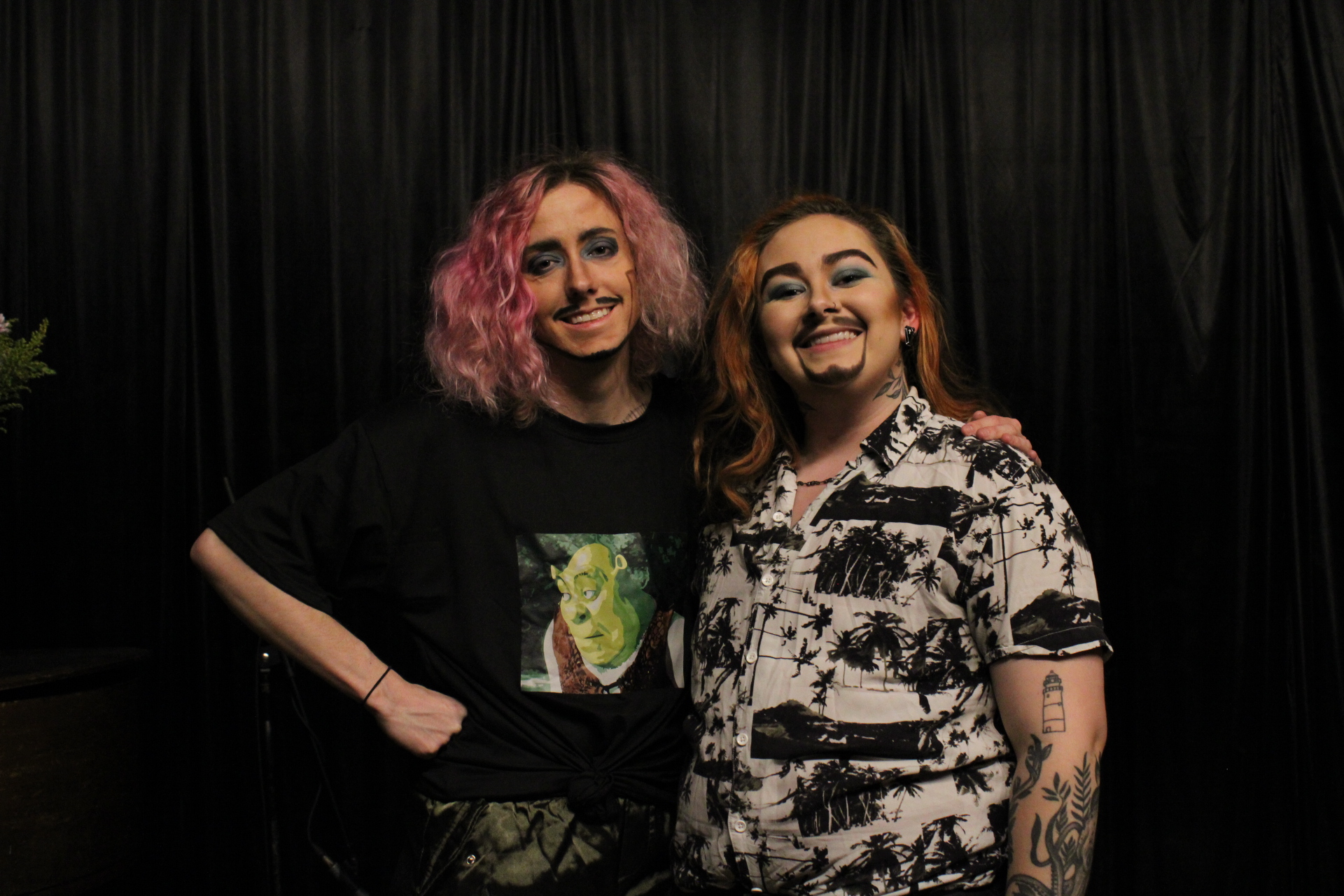 Two smiling women with colorful hair stand close together against a black curtain background.