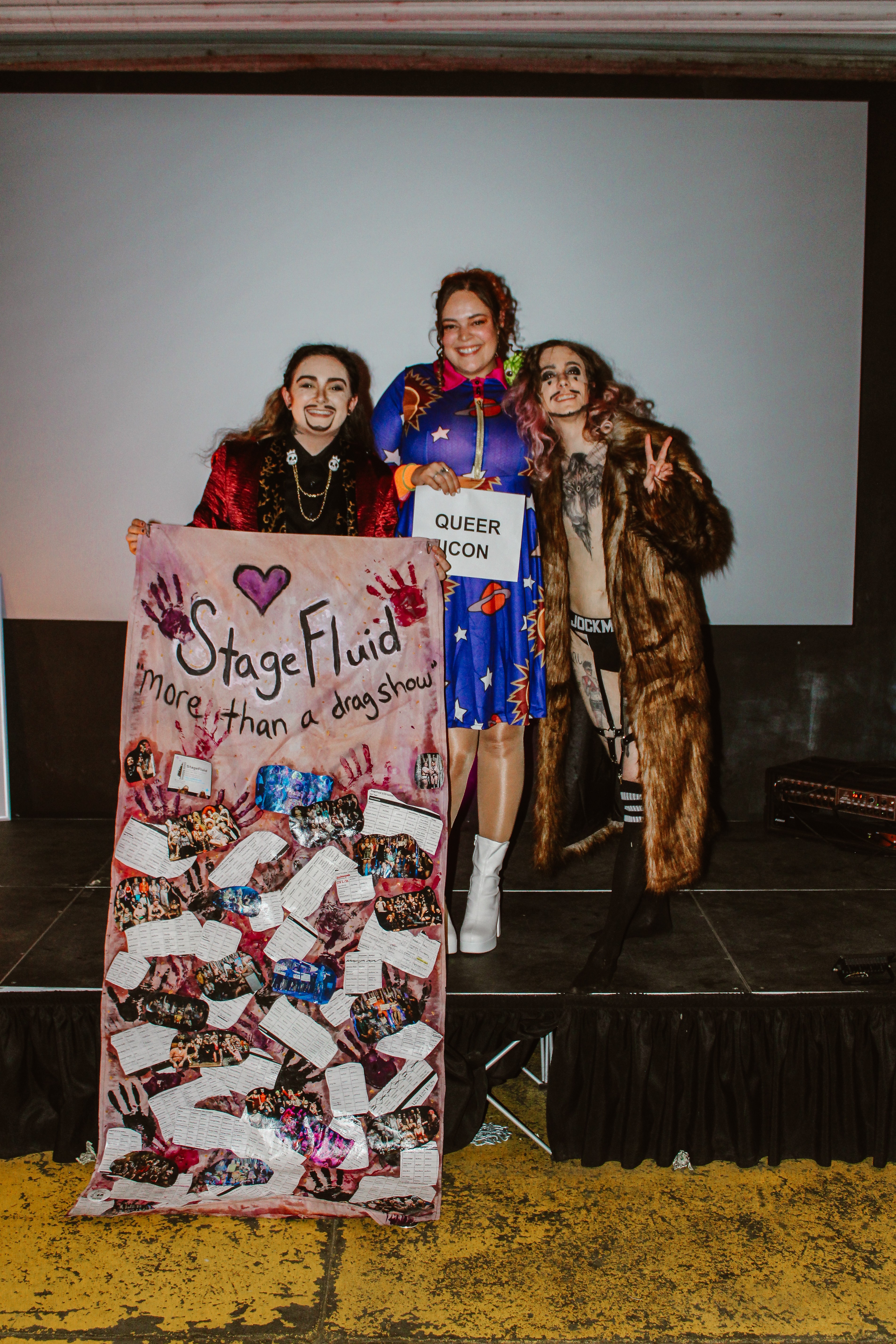 Three people standing on stage holding a colorful sign, with a plain background and a decorated poster in front.