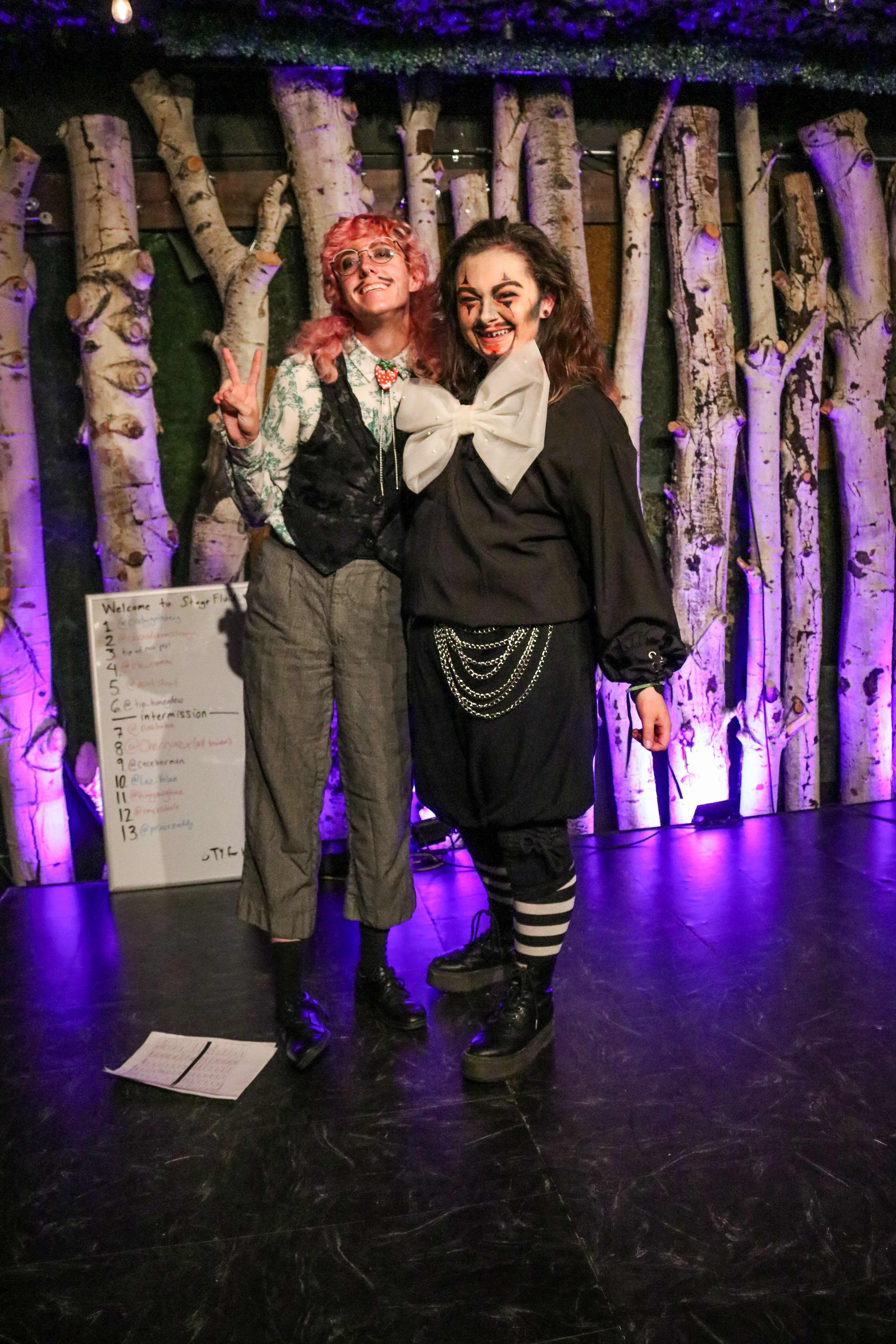 Two people in costumes stand together on stage with a wooden backdrop and purple lighting.