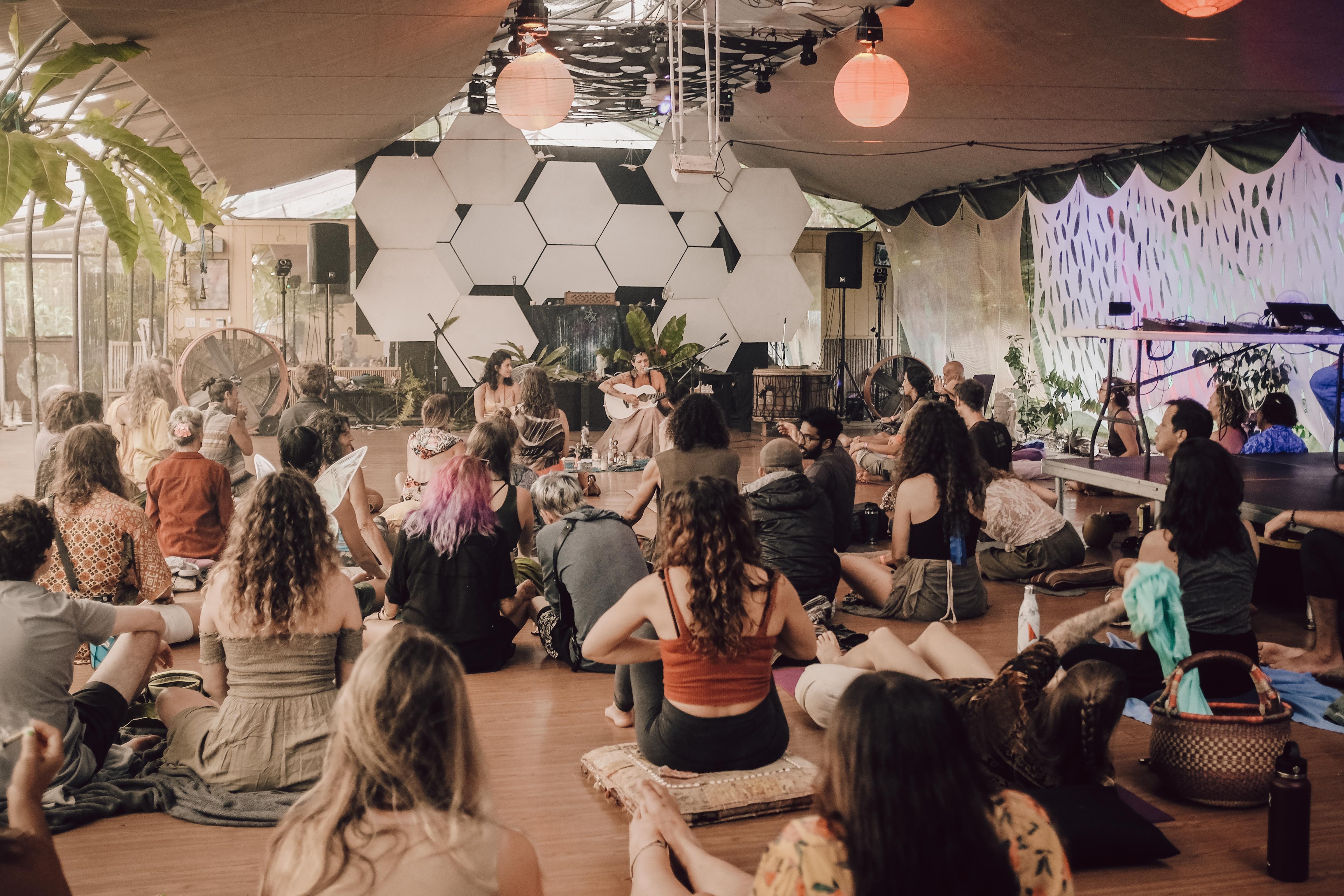 People sitting on the floor facing a stage with hexagonal decorations, in a decorated indoor venue with hanging lights.