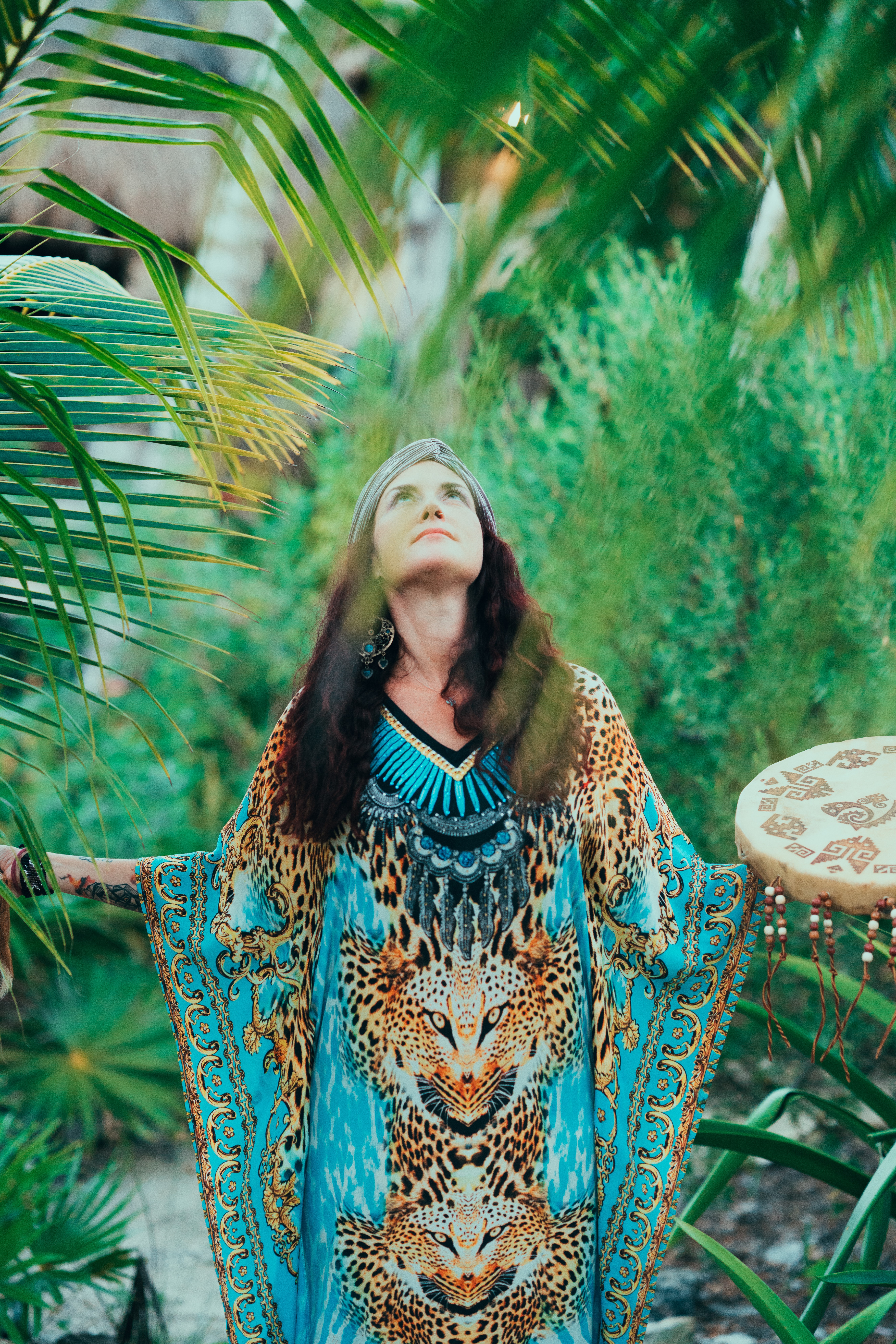 Woman with long dark hair in a colorful animal print dress stands among green plants, looking upward with arms outstretched.