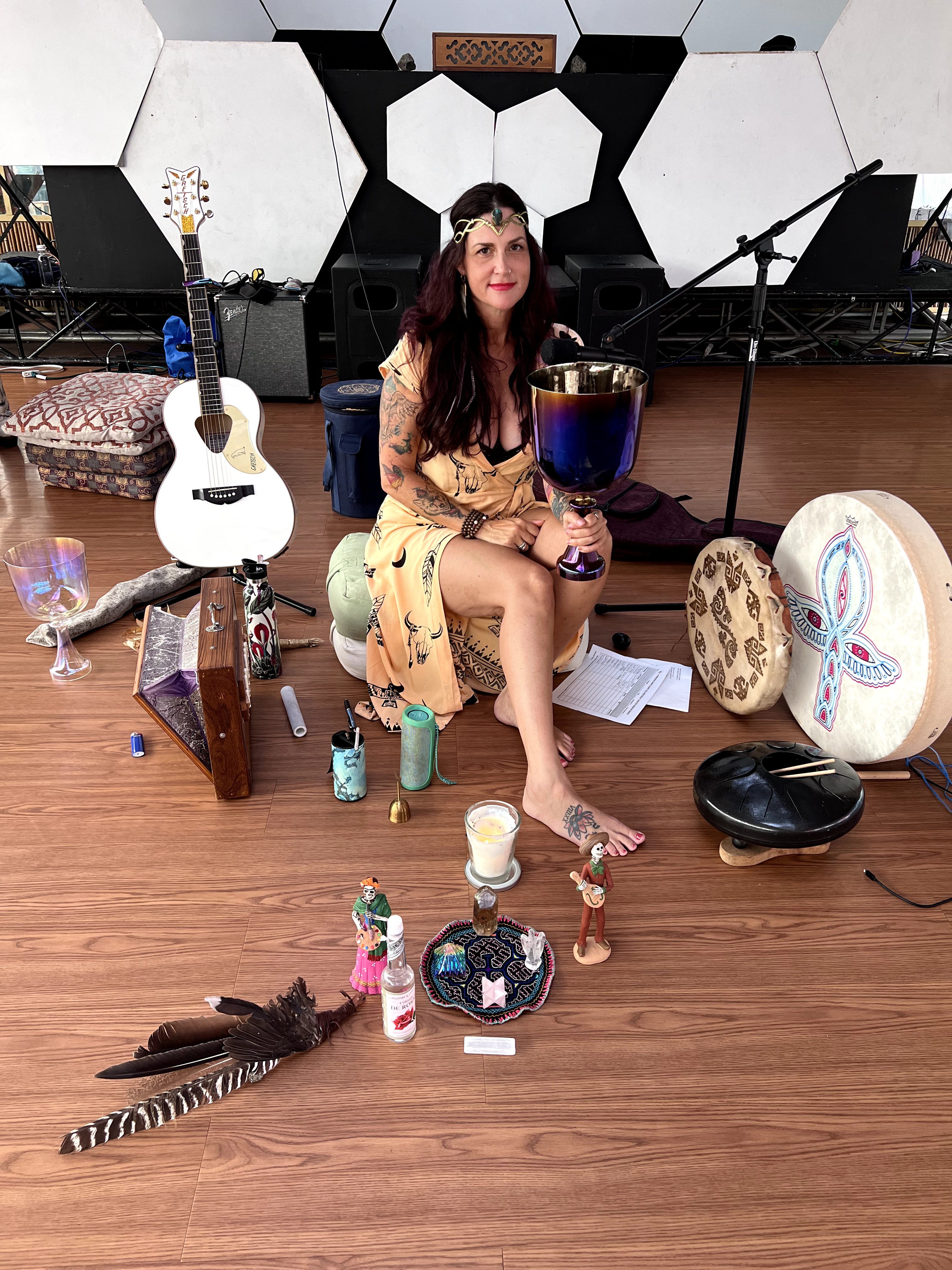 Woman sitting on a stool surrounded by musical instruments and decorations, with a guitar, drums, and feathers on the floor.