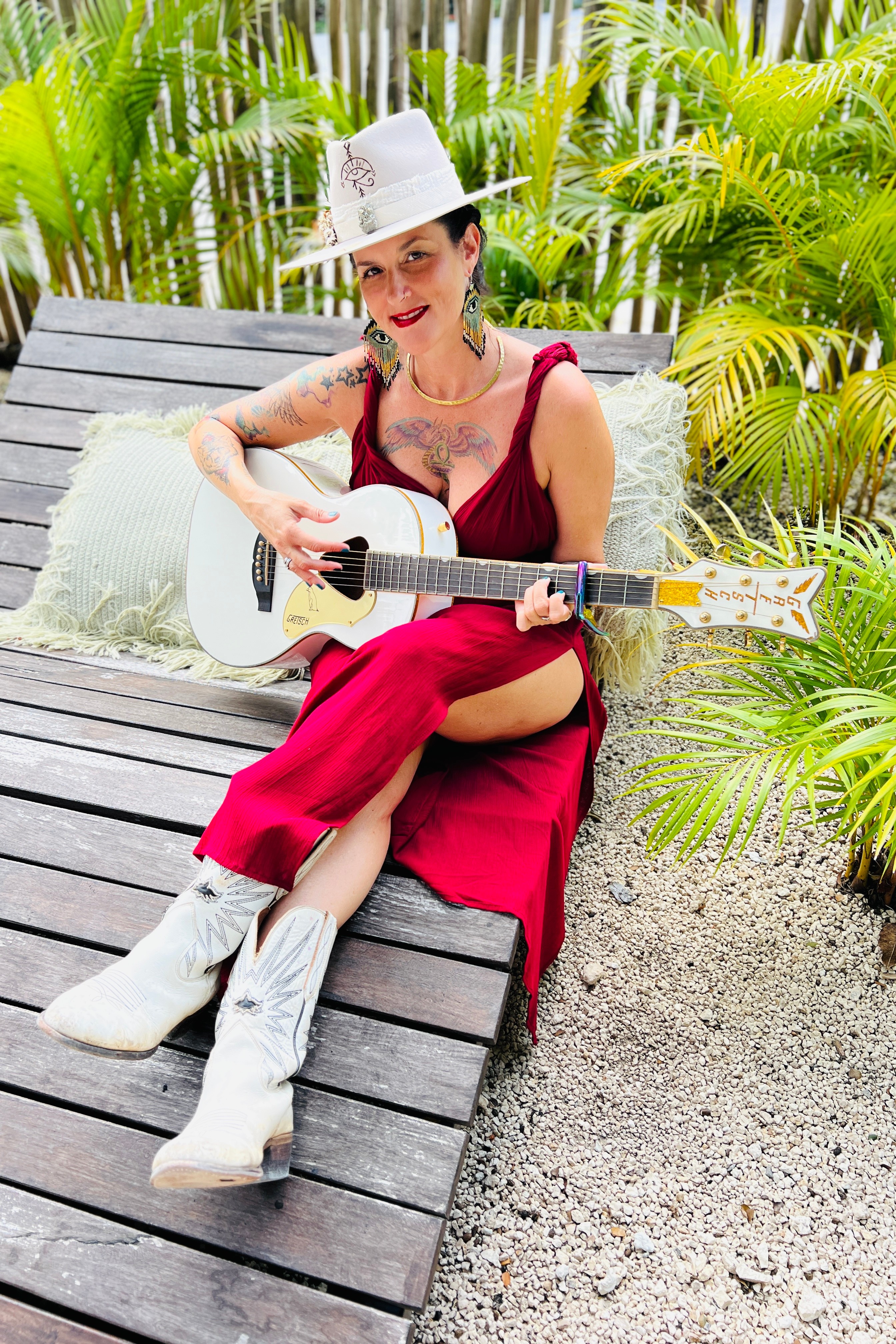 Woman in red dress and white hat sitting on wooden bench playing guitar outdoors surrounded by green plants.