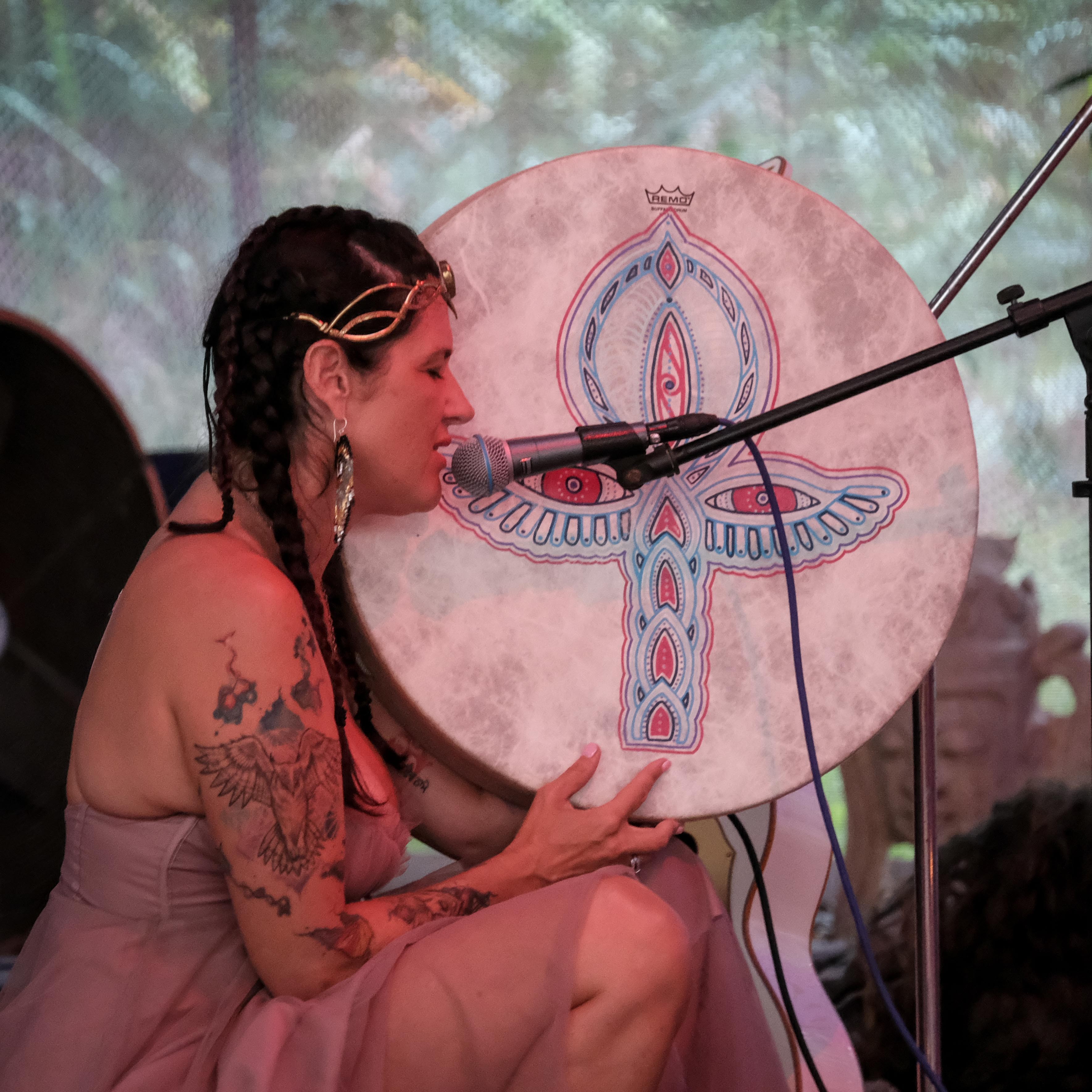 Woman with tattoos and braided hair sings into a microphone, holding a decorated drum with a butterfly design.