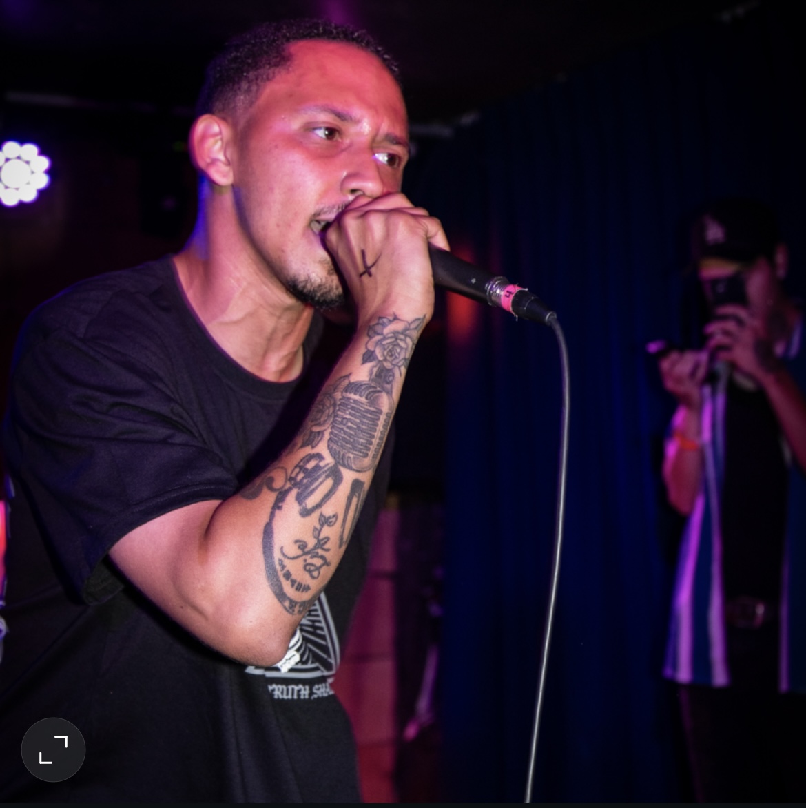 Person singing into a microphone with tattoos on arm, in a dark setting with stage lighting.