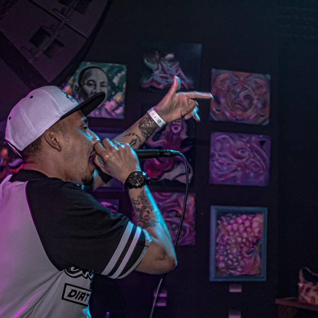A person with tattoos, wearing a cap and a sports jersey, singing into a microphone on stage with colorful artwork behind.