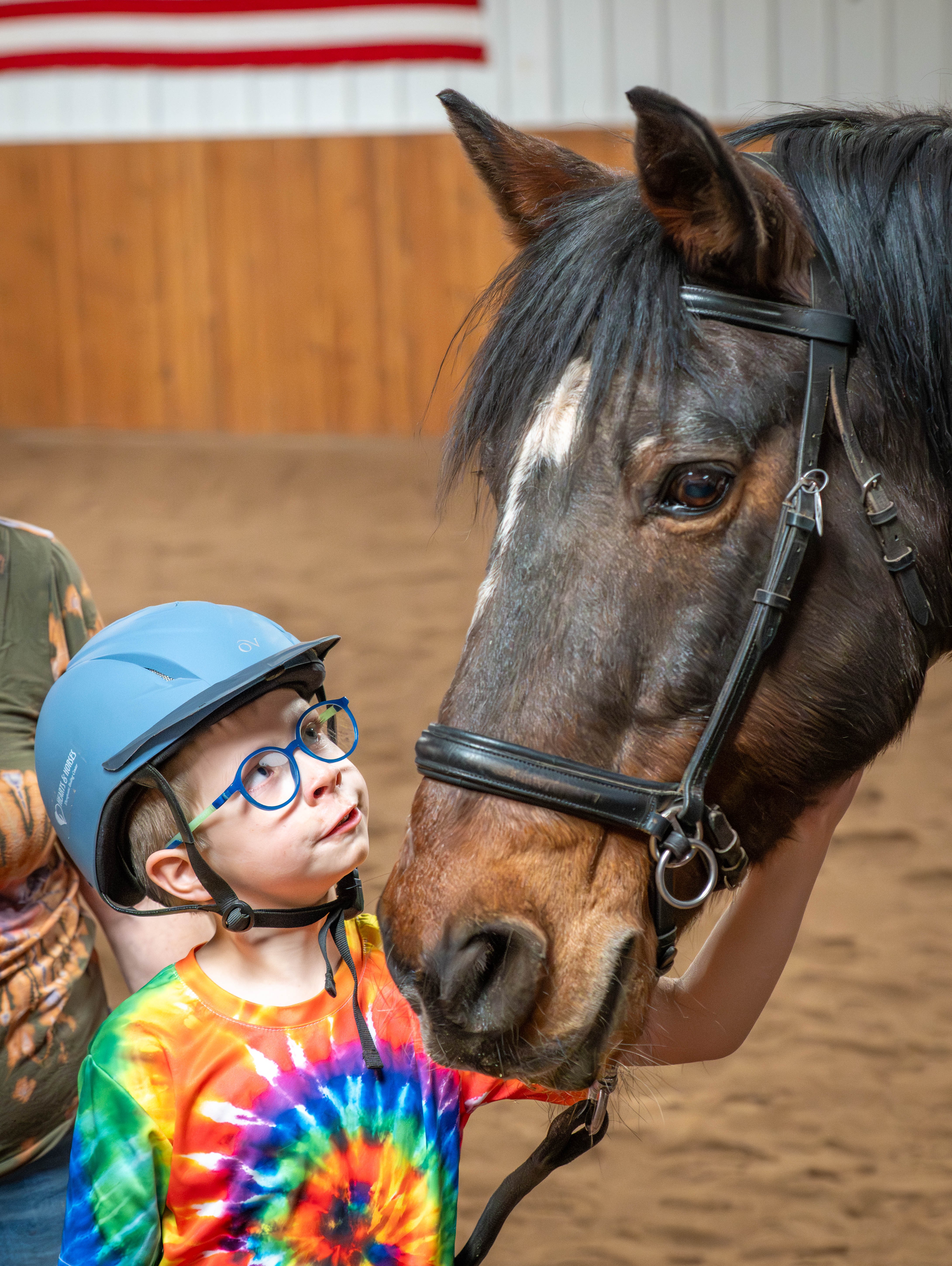 Child wearing glasses and a rainbow tie-dye shirt looks at a horse in an indoor riding arena.