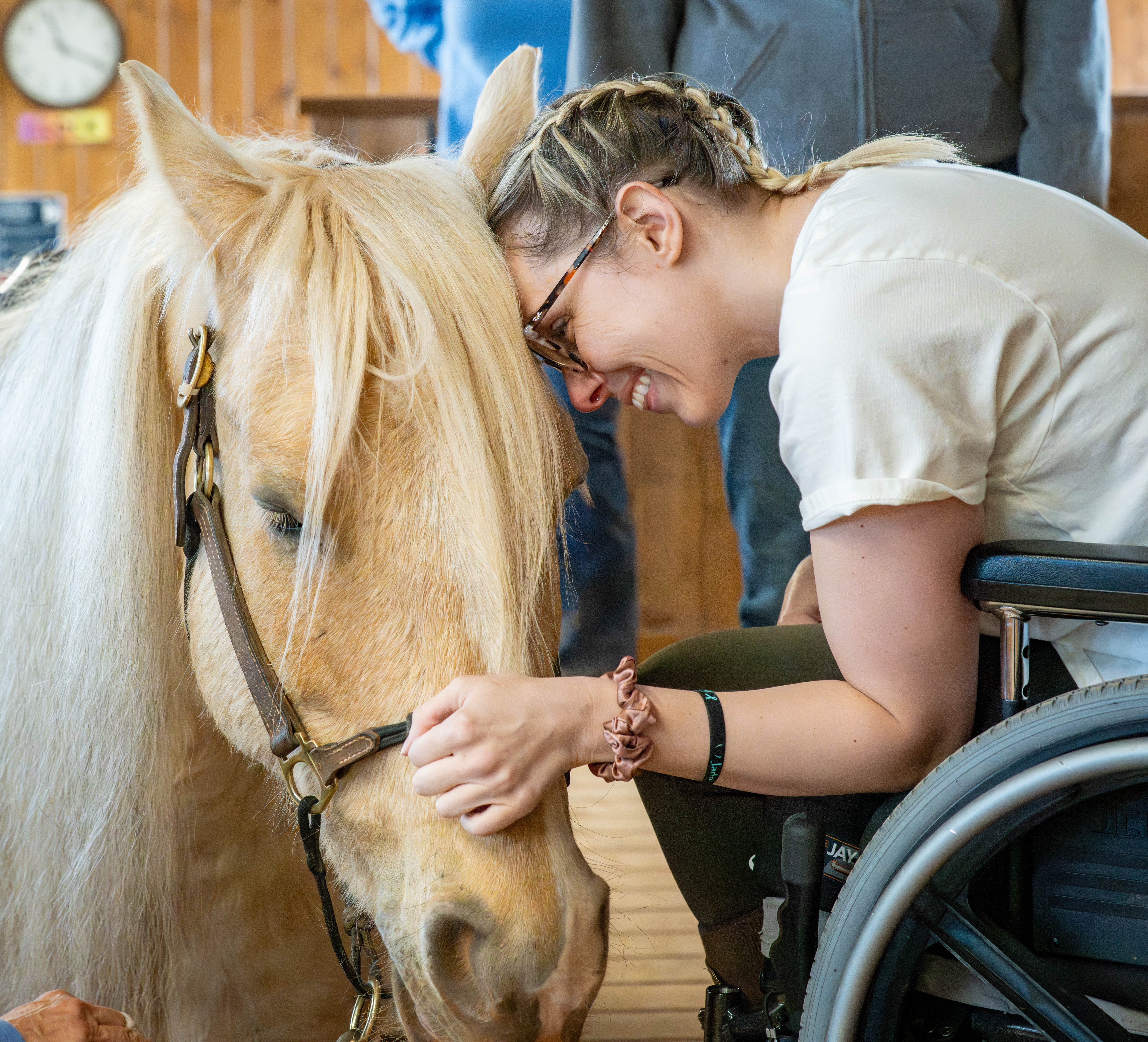 Young woman in a wheelchair gently touches a horse's face, smiling and leaning close indoors.