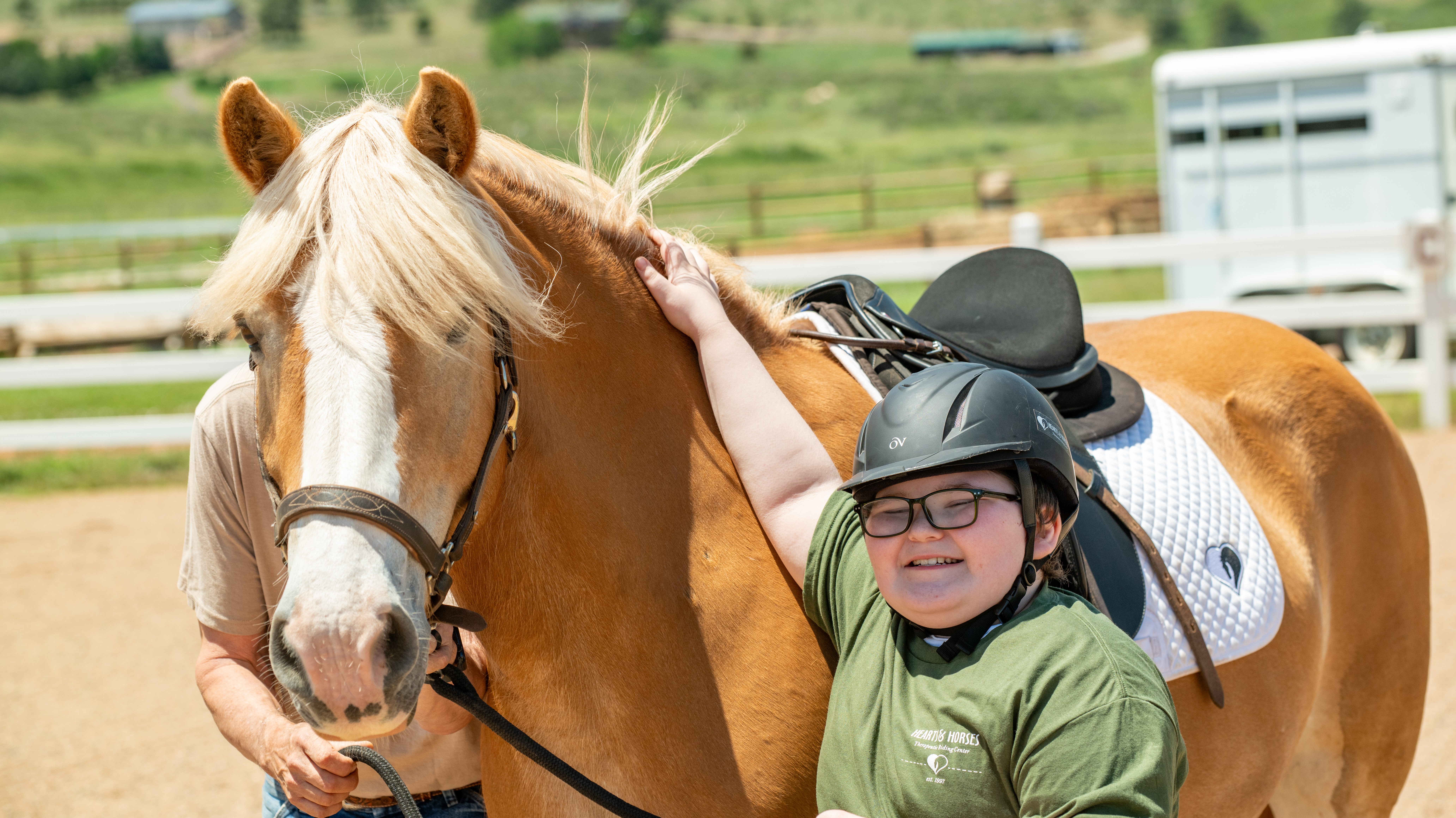 Child wearing helmet petting a horse's head outdoors, with a person holding the horse's bridle nearby.