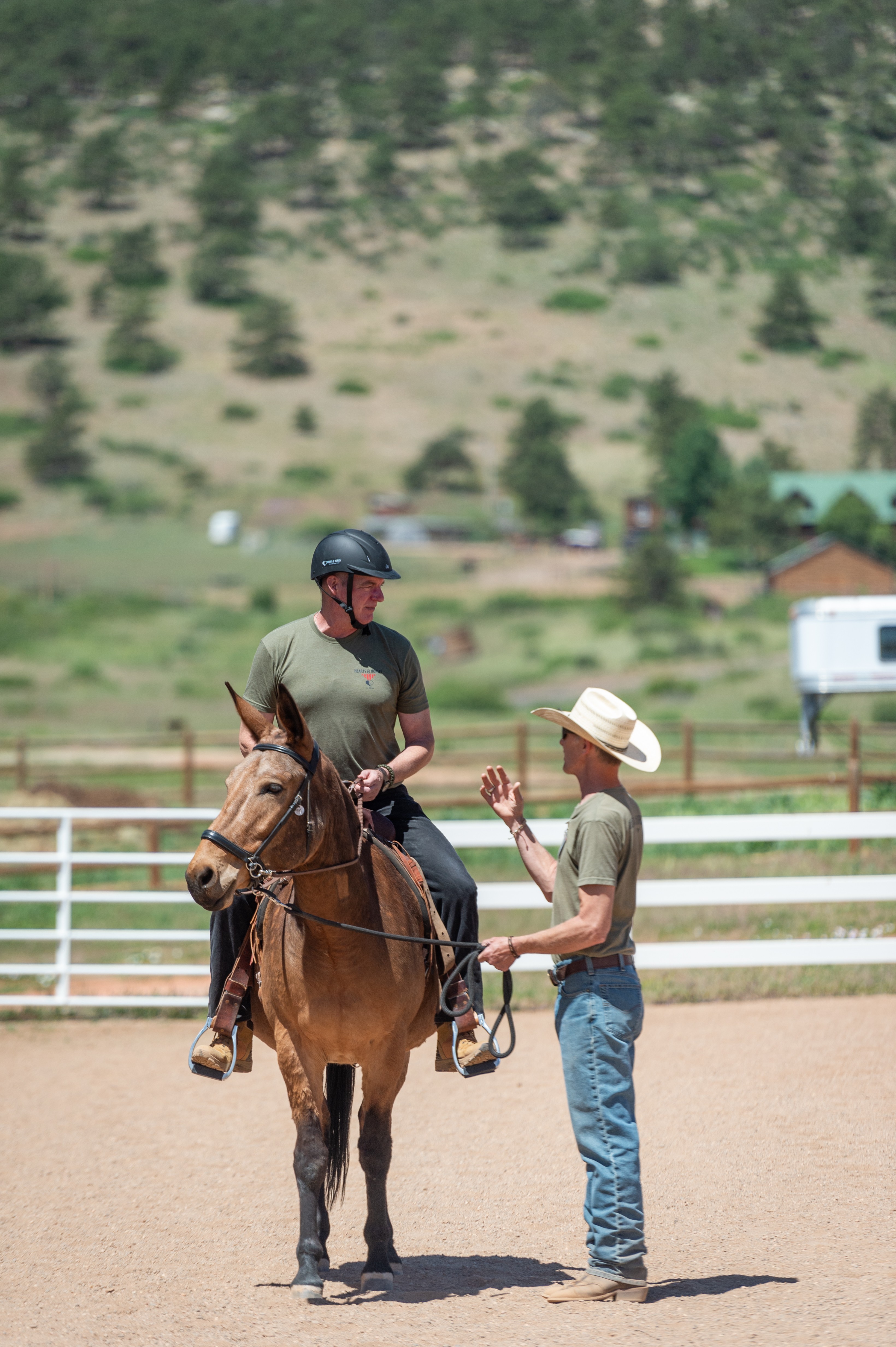 Two women, one on horseback and one standing, converse in an outdoor riding arena with a fence and hills in background.
