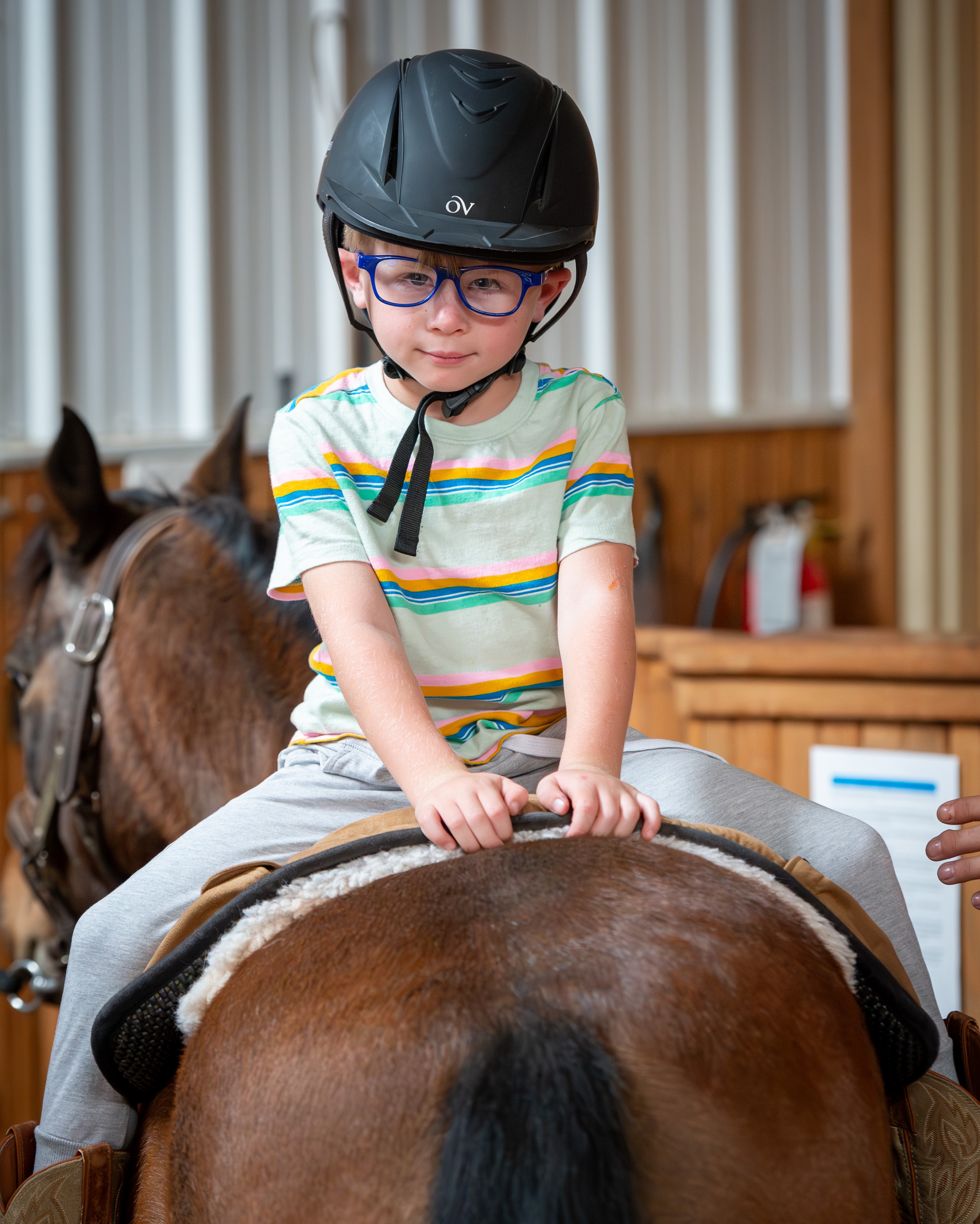 Child wearing glasses and a helmet, sitting on a horse indoors, holding the saddle, with another horse nearby.