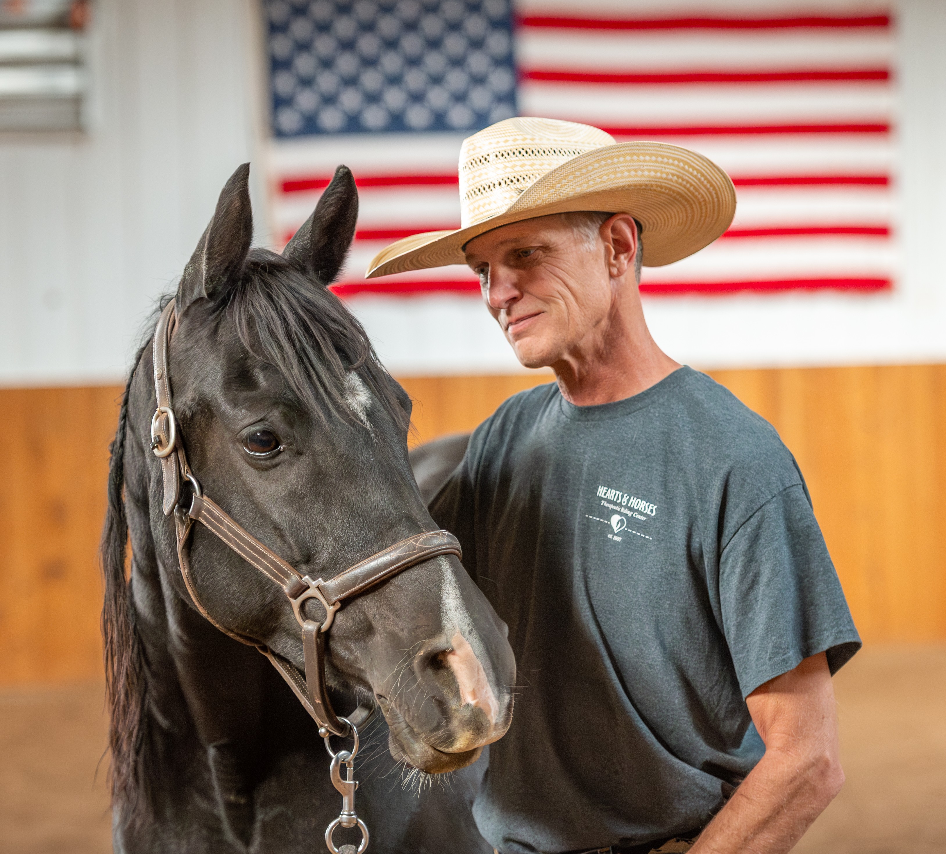 Man wearing a straw hat and gray T-shirt stands next to a black horse inside a barn with an American flag in the background.