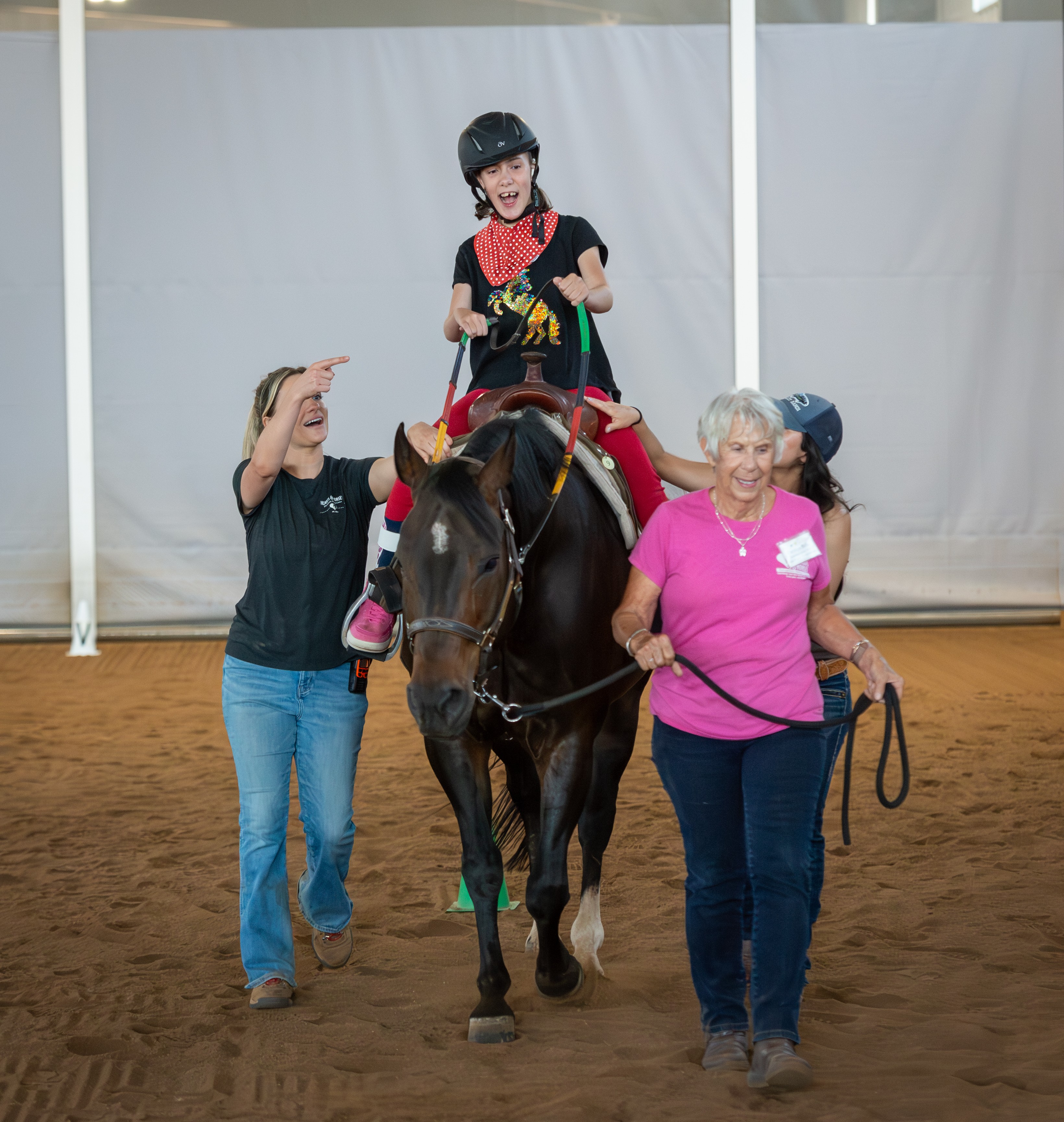 Child in riding gear sits on a horse, guided by two women, indoors on a dirt floor.