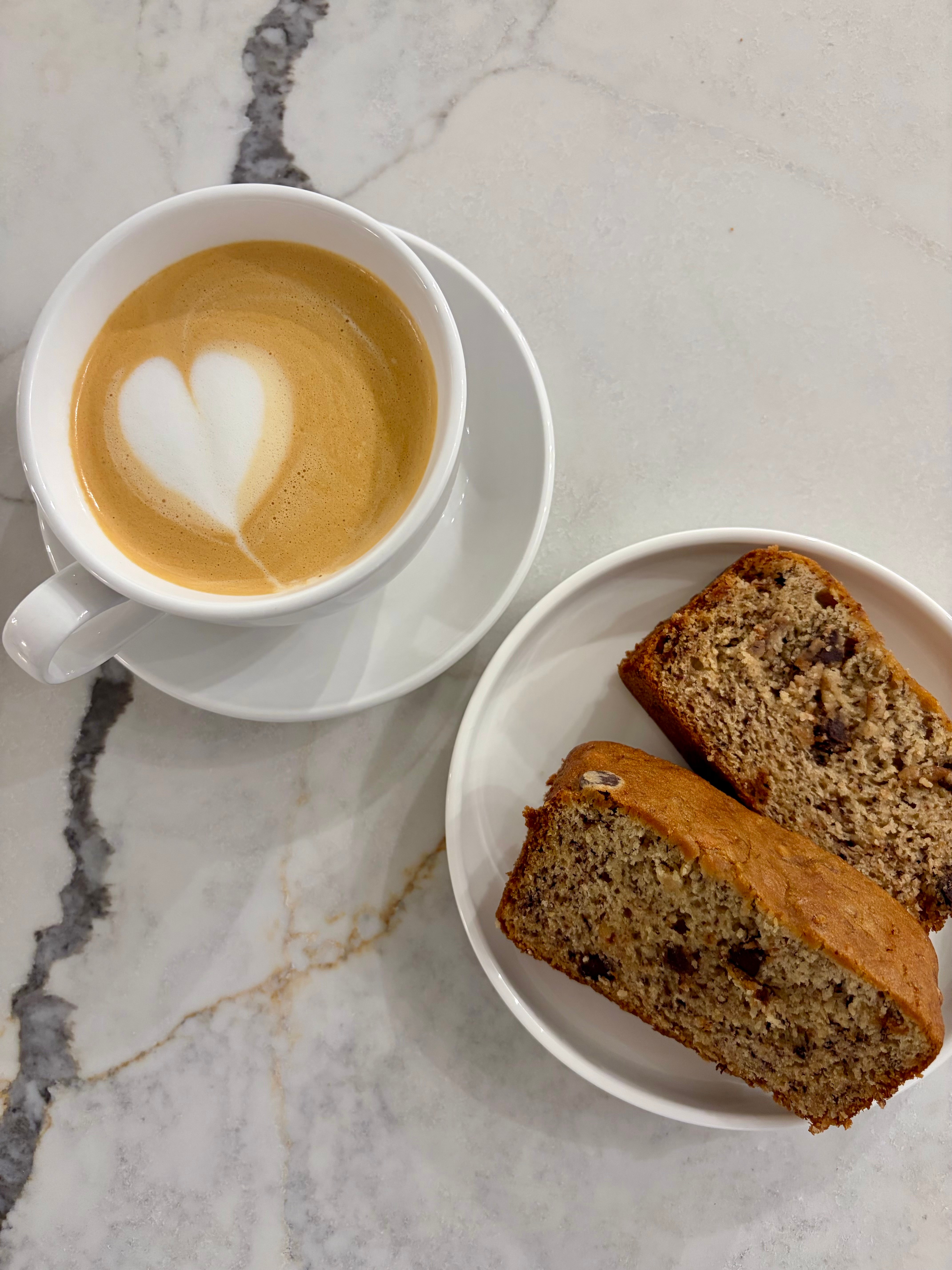 Cup of coffee with heart-shaped foam design and two slices of banana bread on a white plate on a marble surface.