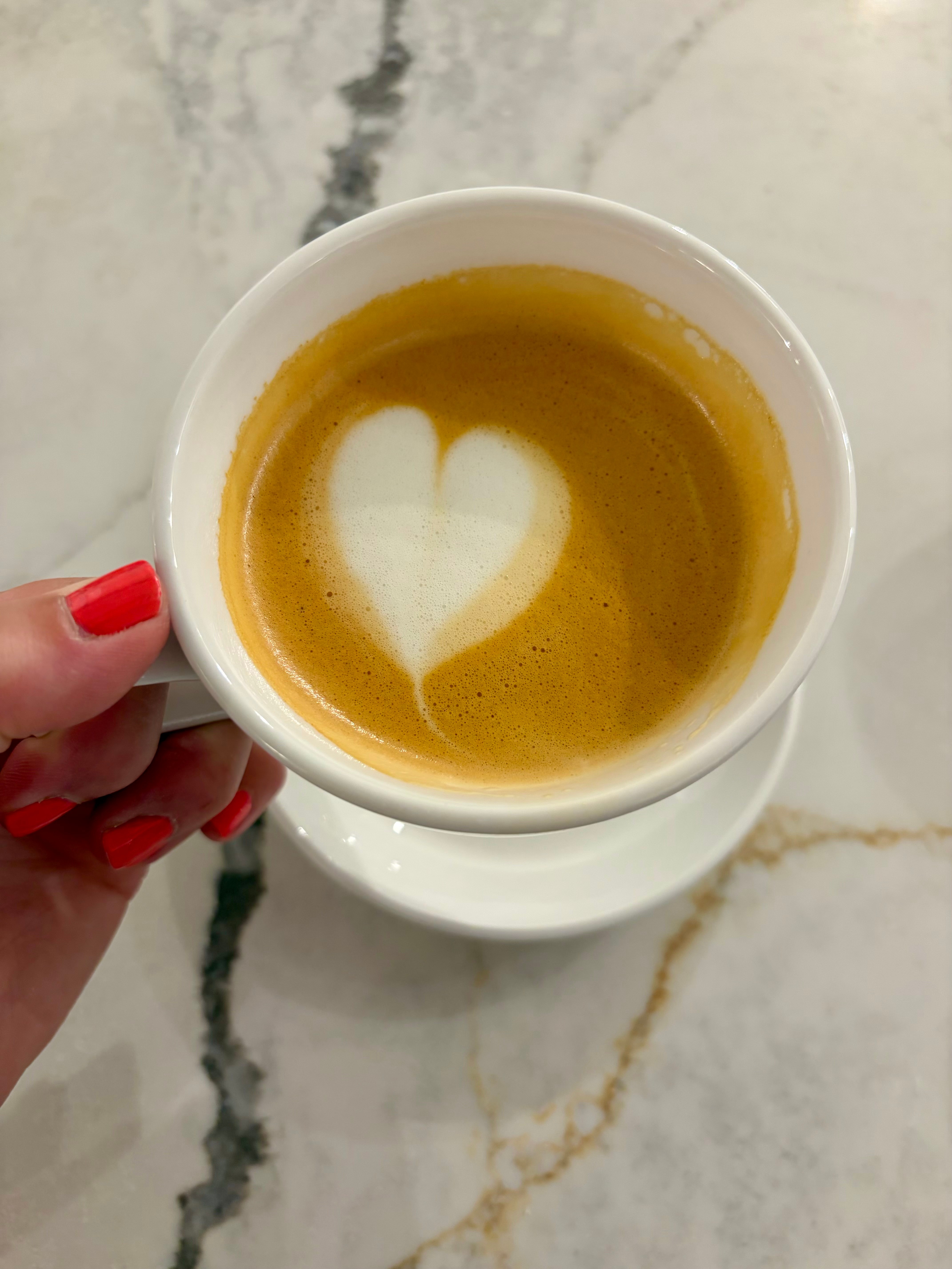 White cup of coffee with heart-shaped foam design, held by a person with red nail polish, on a marble surface.