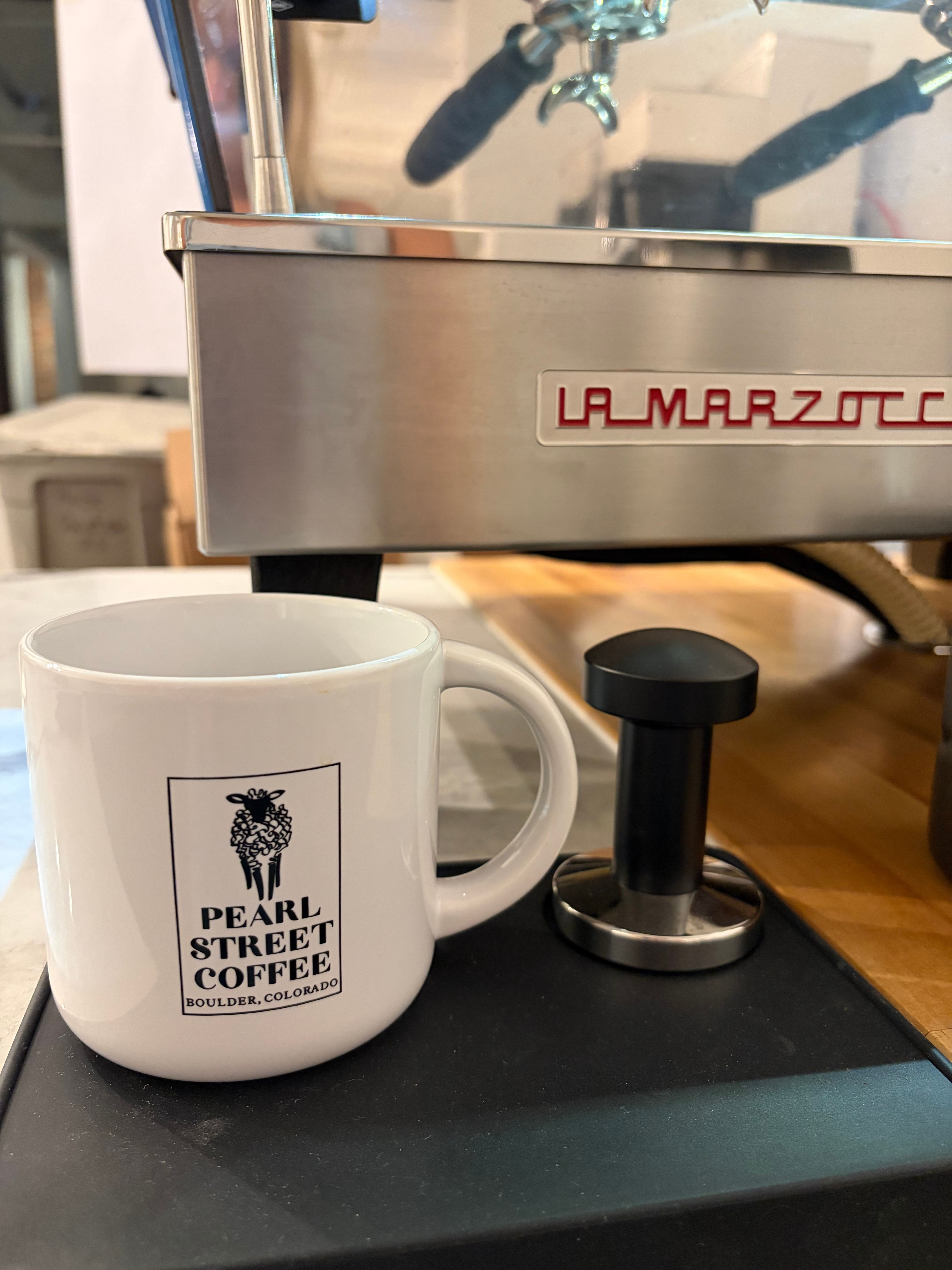 White coffee mug with logo and text, placed on a black surface, with a machine and a handle nearby.
