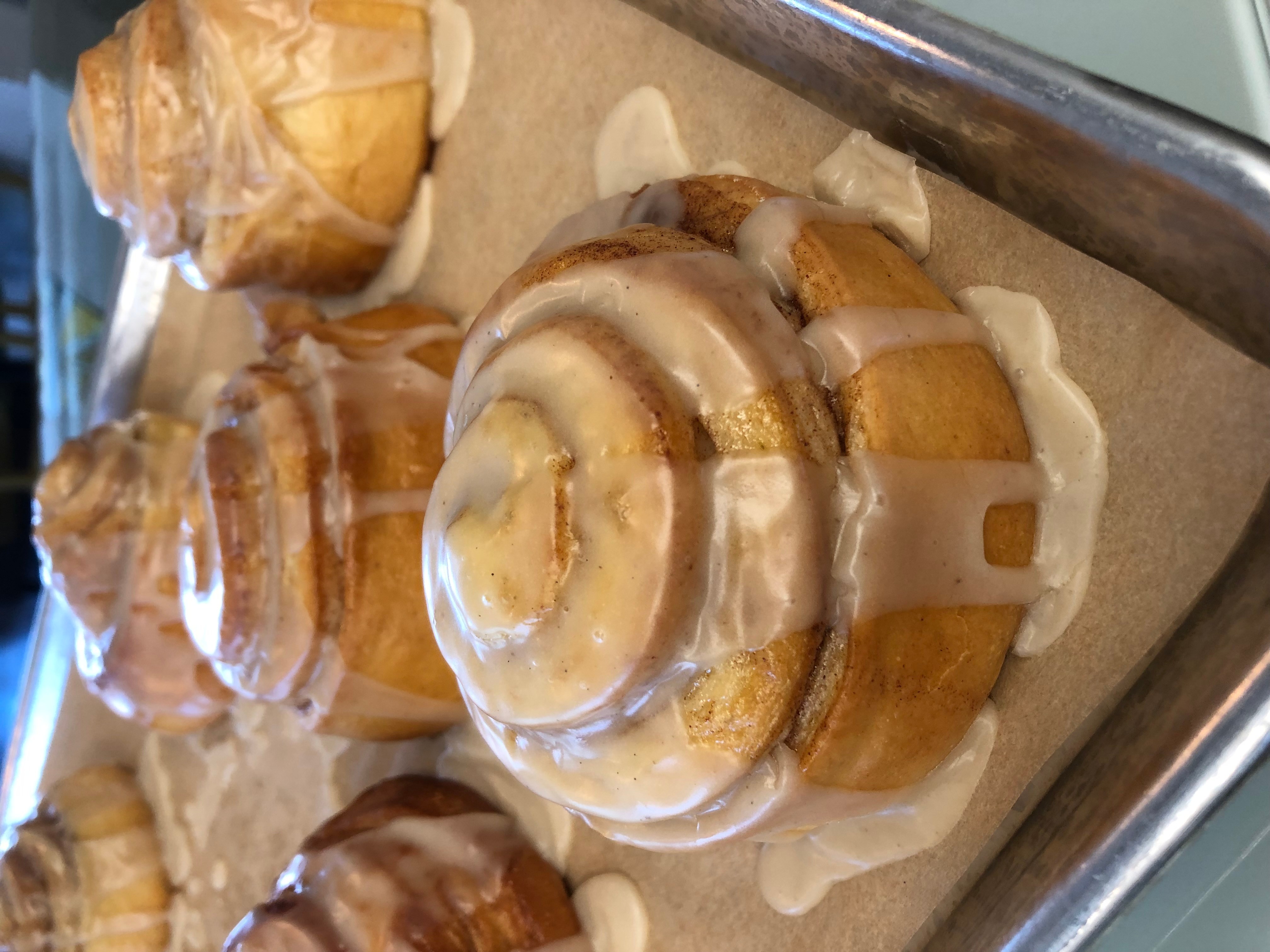 Cinnamon rolls with icing on a baking sheet, some icing melting and dripping, with a pastry in the background.