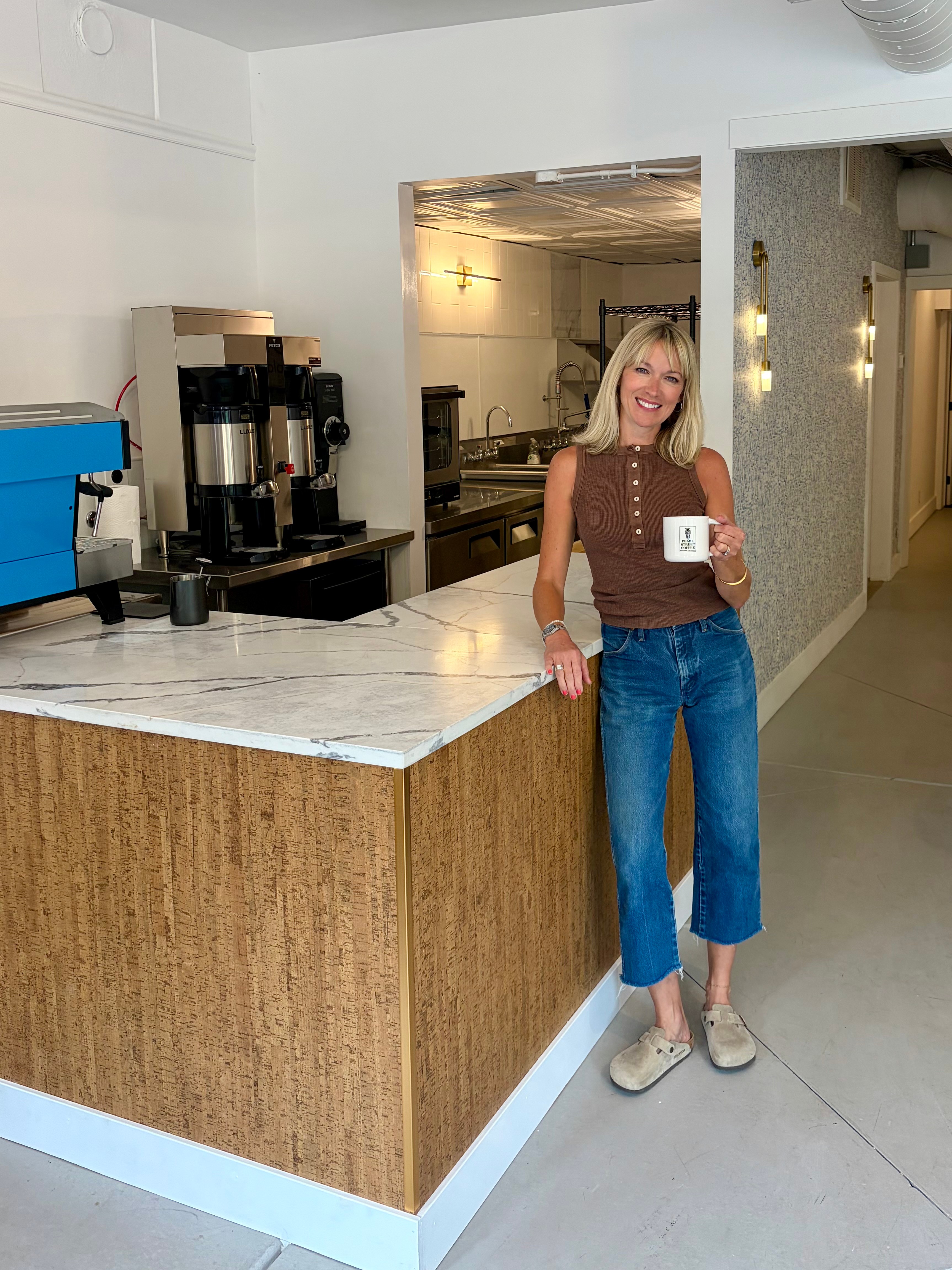 Woman with blonde hair, wearing a brown sleeveless top and blue jeans, standing next to a marble counter in a room.