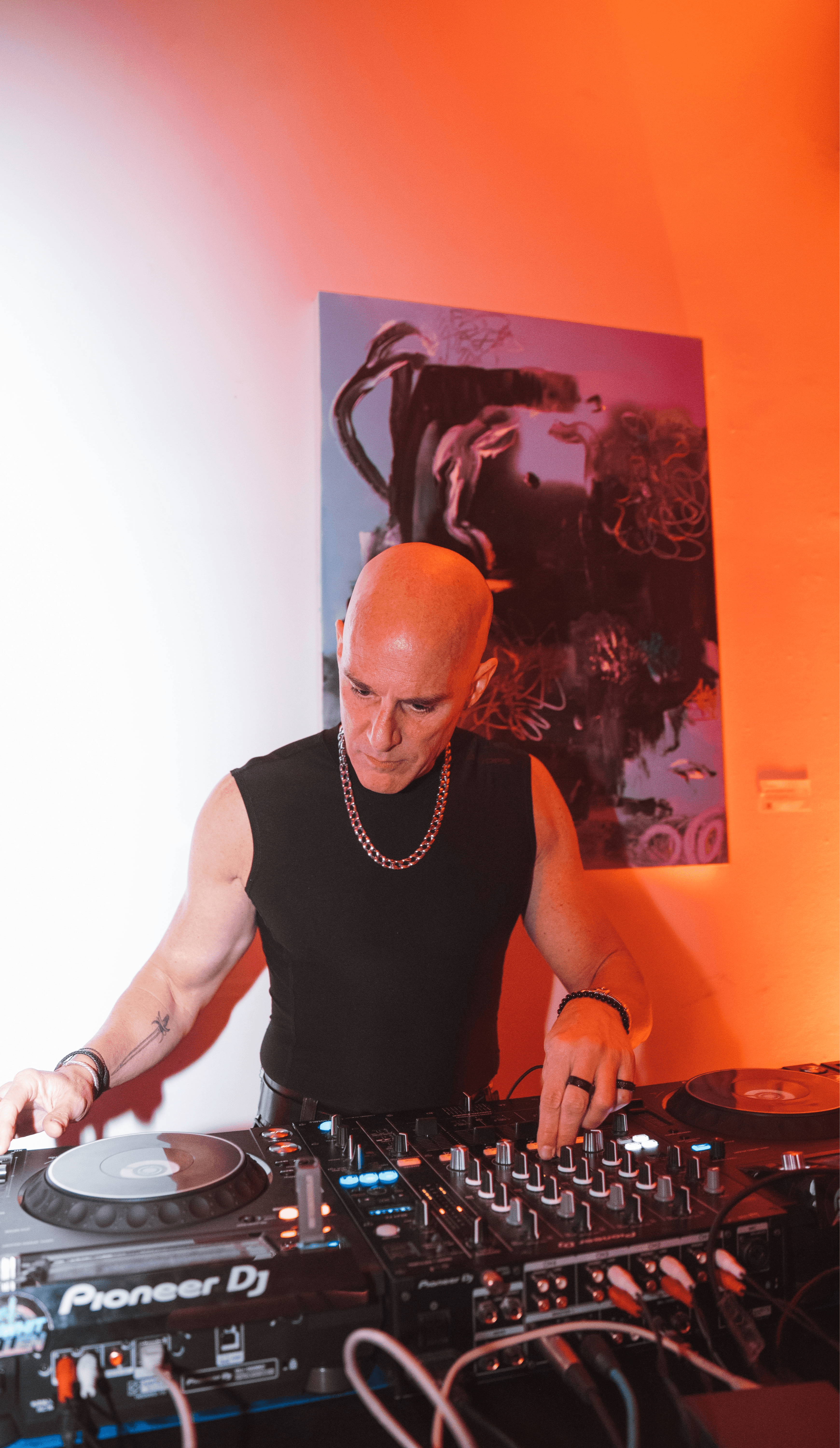 DJ with a bald head and black sleeveless shirt operates a Pioneer DJ controller in a room with orange and white walls, and a colorful painting.