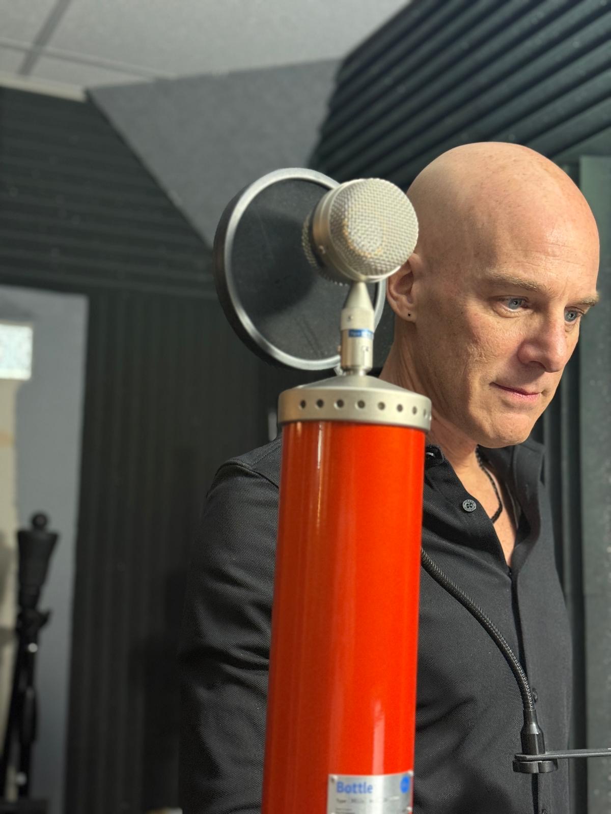 Bald man in black shirt speaking into a large red and silver microphone in a recording studio.