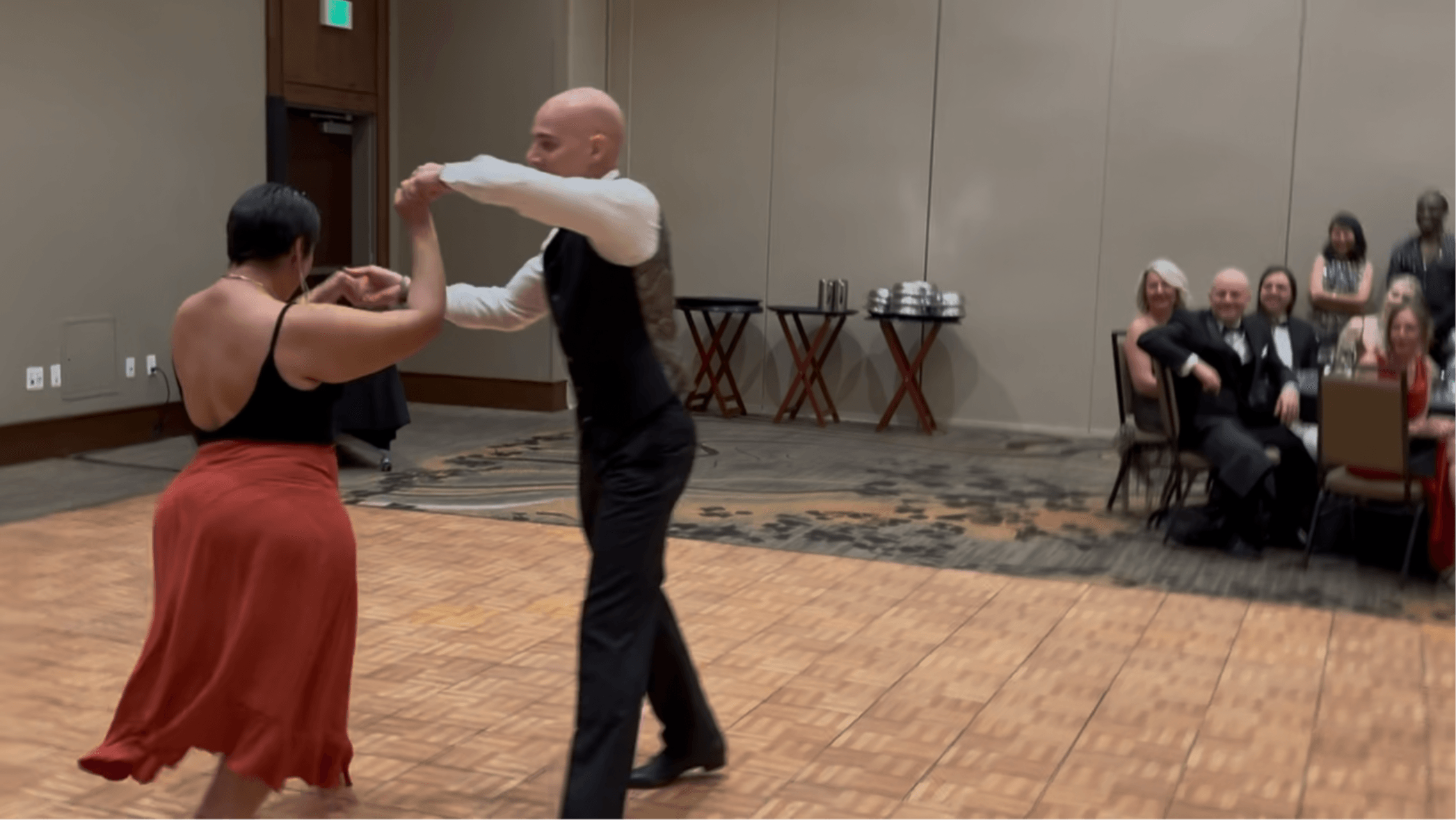 Two people dancing on a wooden floor with an audience seated in the background.
